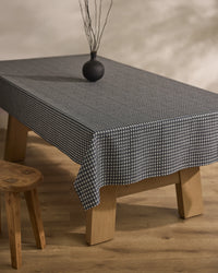 A rectangular wooden table is draped with Geometry’s Houndstooth Black tablecloth, topped with a small round black vase holding dry branches. A wooden stool sits beside the table on a light wooden floor.