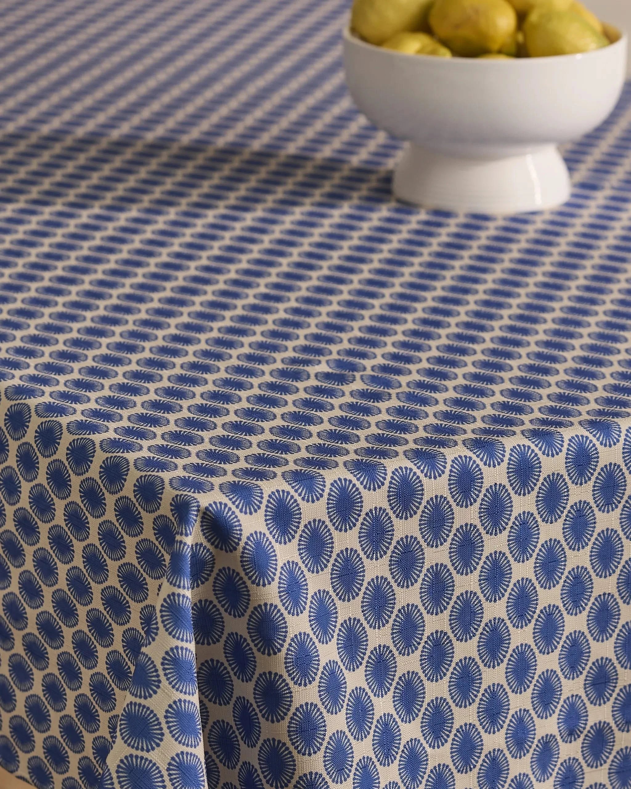 A table is draped with the Geometry Lagoon stain-resistant tablecloth, showcasing blue and white circular patterns, and a white bowl of pears sits near the top right corner.