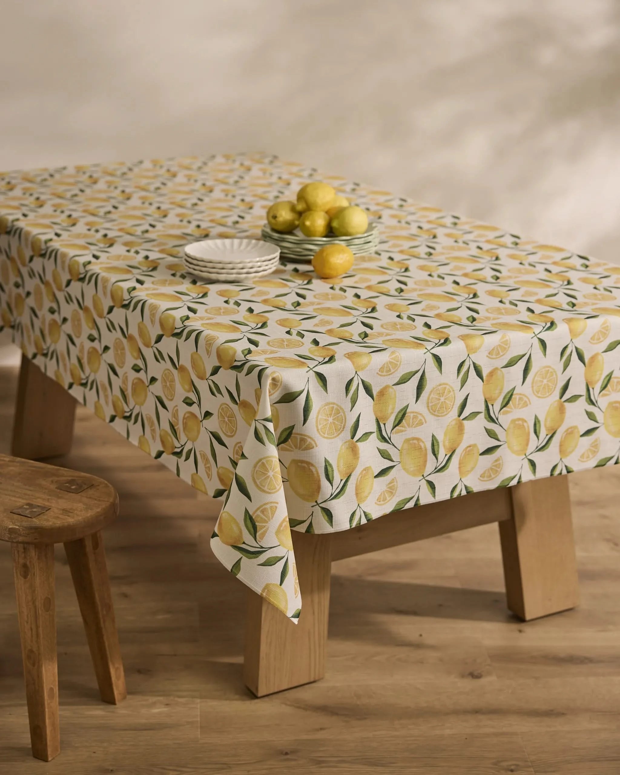 A wooden table draped with the Geometry Limoncello stain-resistant tablecloth displays white plates and a bowl of lemons; a wooden stool rests nearby on the light wood floor.