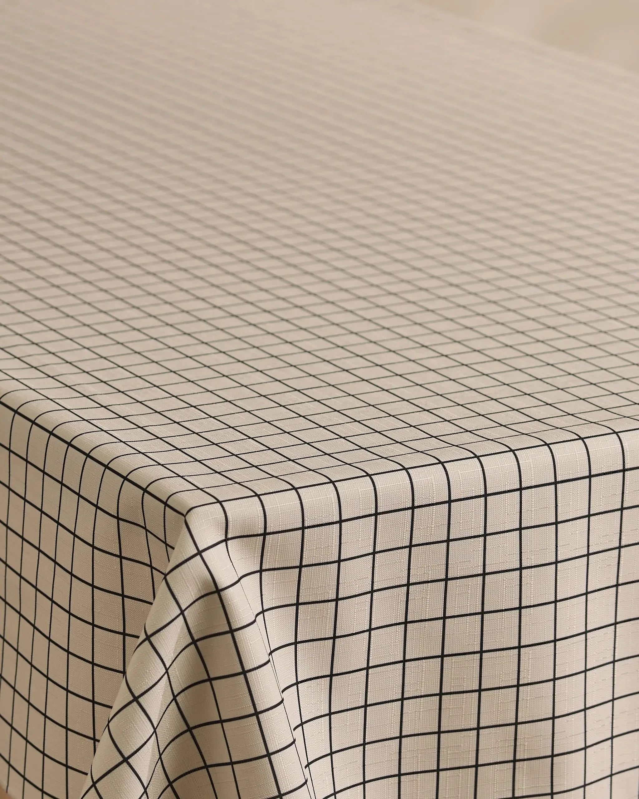 The Geometry Metro Grid tablecloth features a beige, stain-resistant fabric with evenly spaced black grid lines, draping neatly over rectangular tables for a clean, minimalistic look.