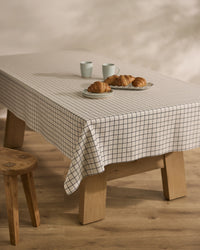 A wooden table with Geometrys Metro Grid stain-resistant tablecloth in a white and black pattern holds four croissants on a plate and two white mugs. A wooden stool is beside the table on a light wooden floor.