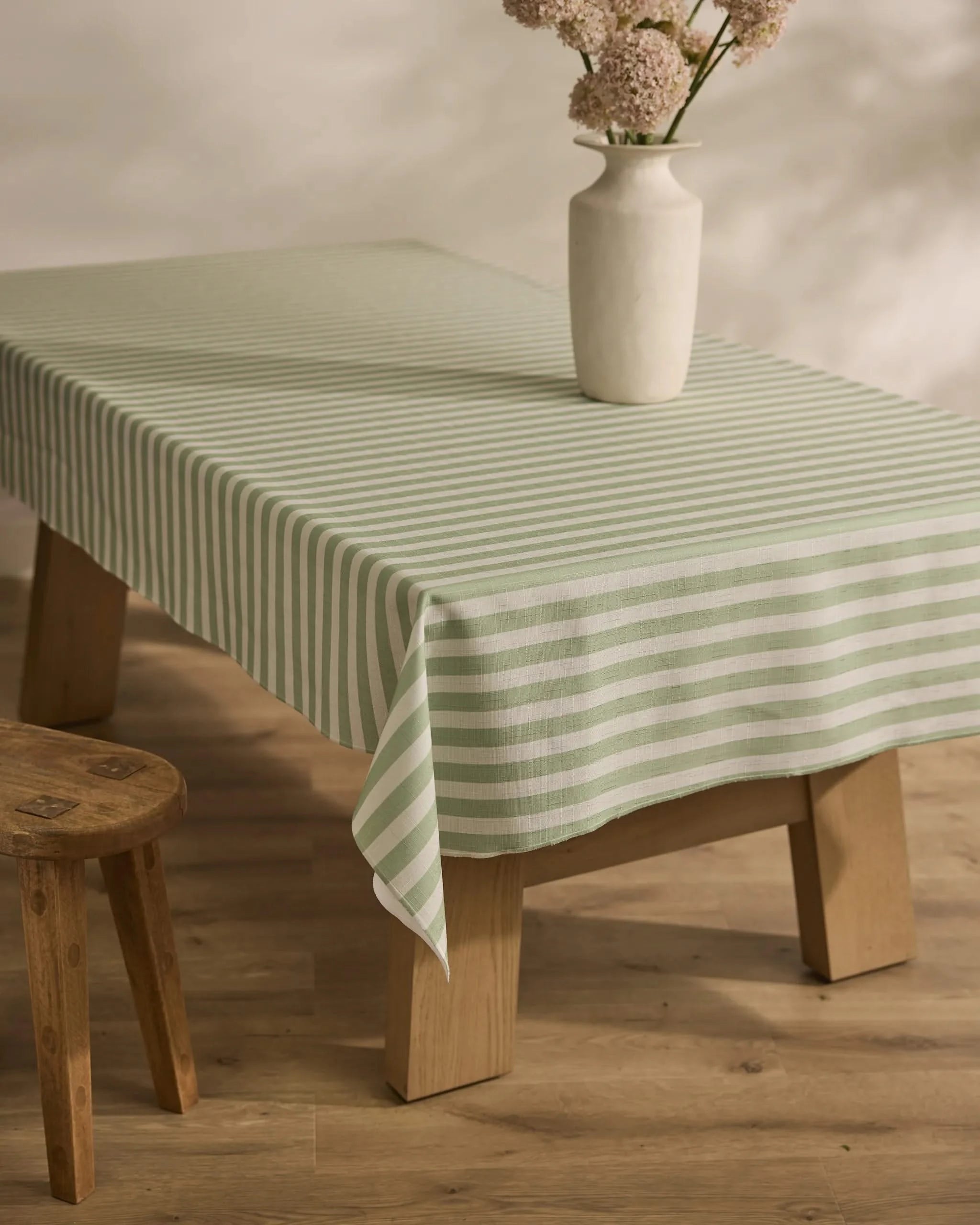 A light wooden table covered with Geometry’s Seagrass green and white stain-resistant tablecloth holds a white vase with pink flowers; a wooden stool sits nearby on the light wooden floor.