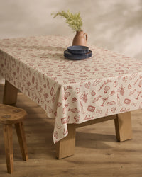 A wooden table is draped with Geometrys Summer Air Tablecloth, featuring red food-themed doodles and Italian words, topped with blue plates and a pink vase of greenery. A wooden stool sits nearby on the wood floor.