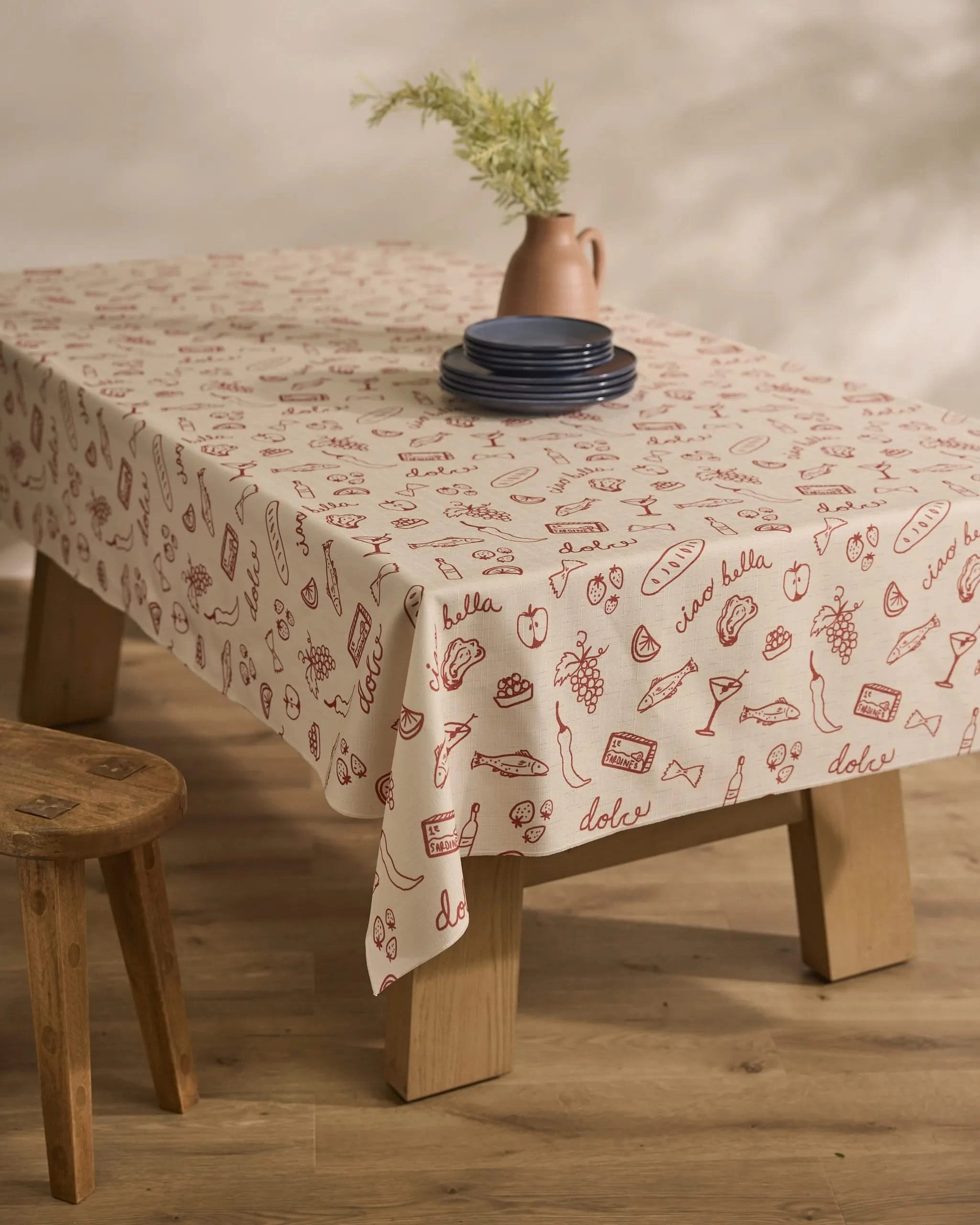 A wooden table is draped with Geometrys Summer Air Tablecloth, featuring red food-themed doodles and Italian words, topped with blue plates and a pink vase of greenery. A wooden stool sits nearby on the wood floor.