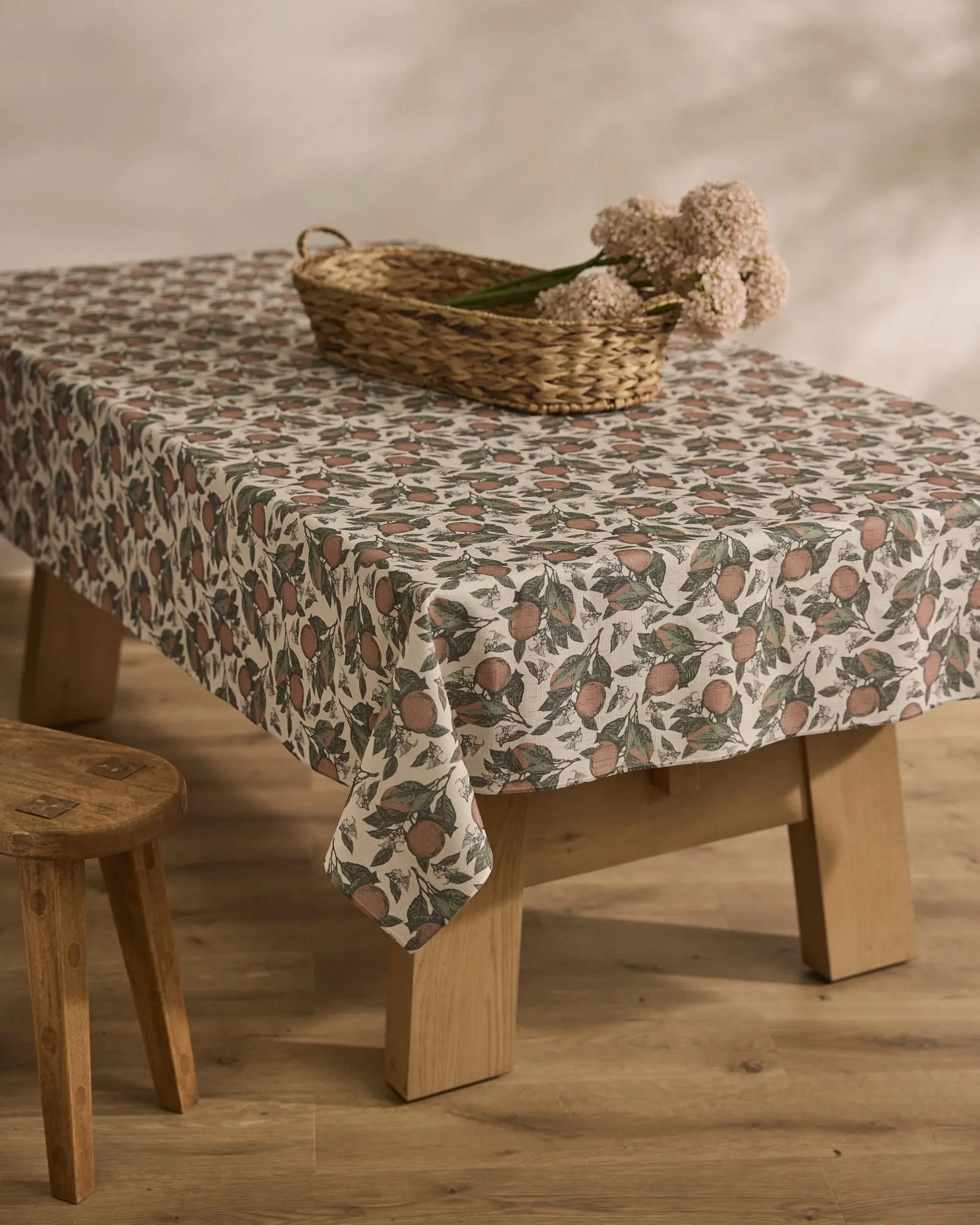 The Valencia tablecloth by Geometry, featuring pomegranates and leaves, covers a wooden table; on top sits a wicker basket with pale pink flowers, while a wooden stool stands beside the table on a wooden floor.