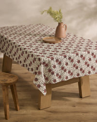 A wooden table is draped with Geometrys Wild Dahlia, a stain-resistant tablecloth with a red floral motif. A brown ceramic plate and matching jug of green foliage sit atop, with a wooden stool placed nearby.