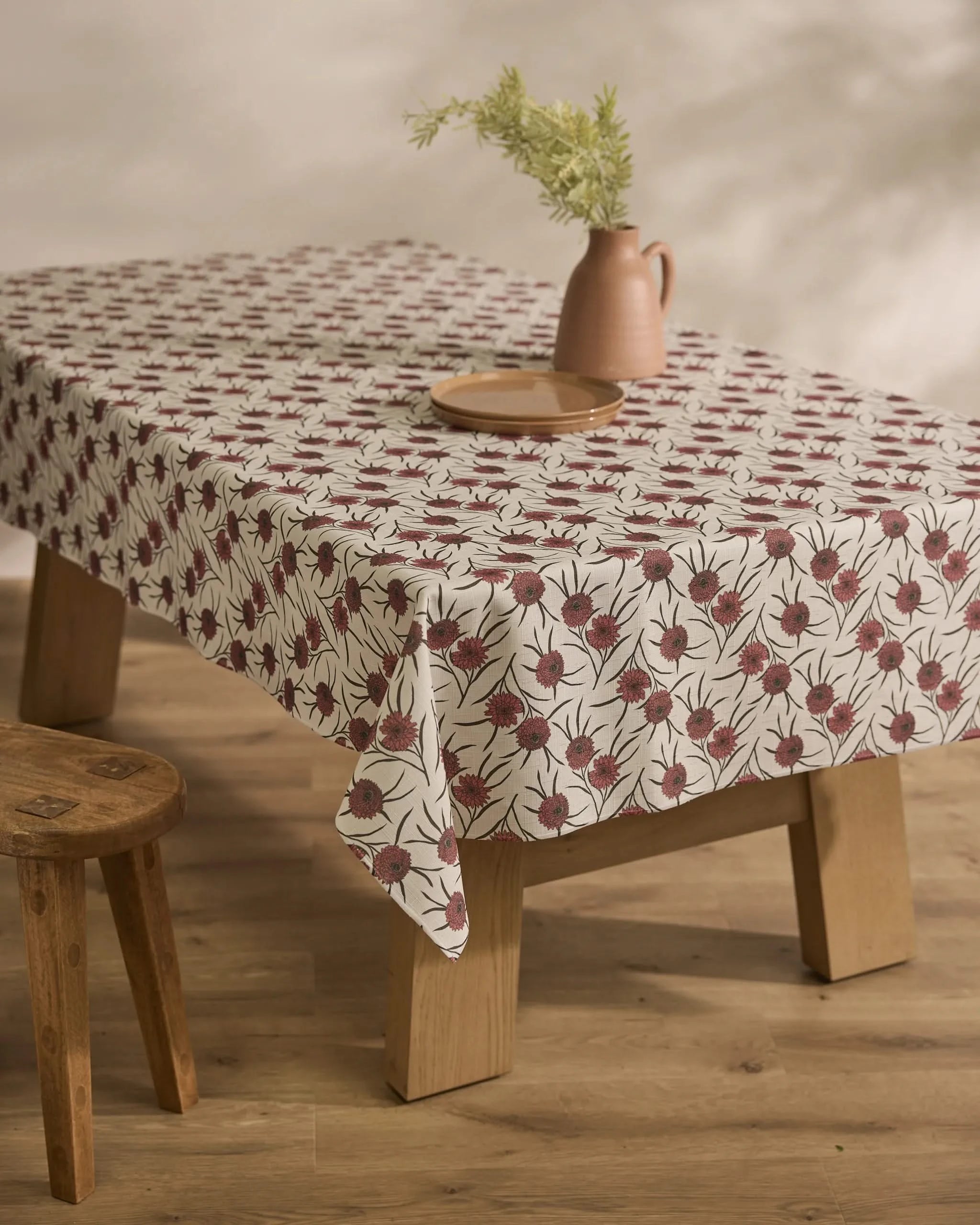 A wooden table is draped with Geometrys Wild Dahlia, a stain-resistant tablecloth with a red floral motif. A brown ceramic plate and matching jug of green foliage sit atop, with a wooden stool placed nearby.