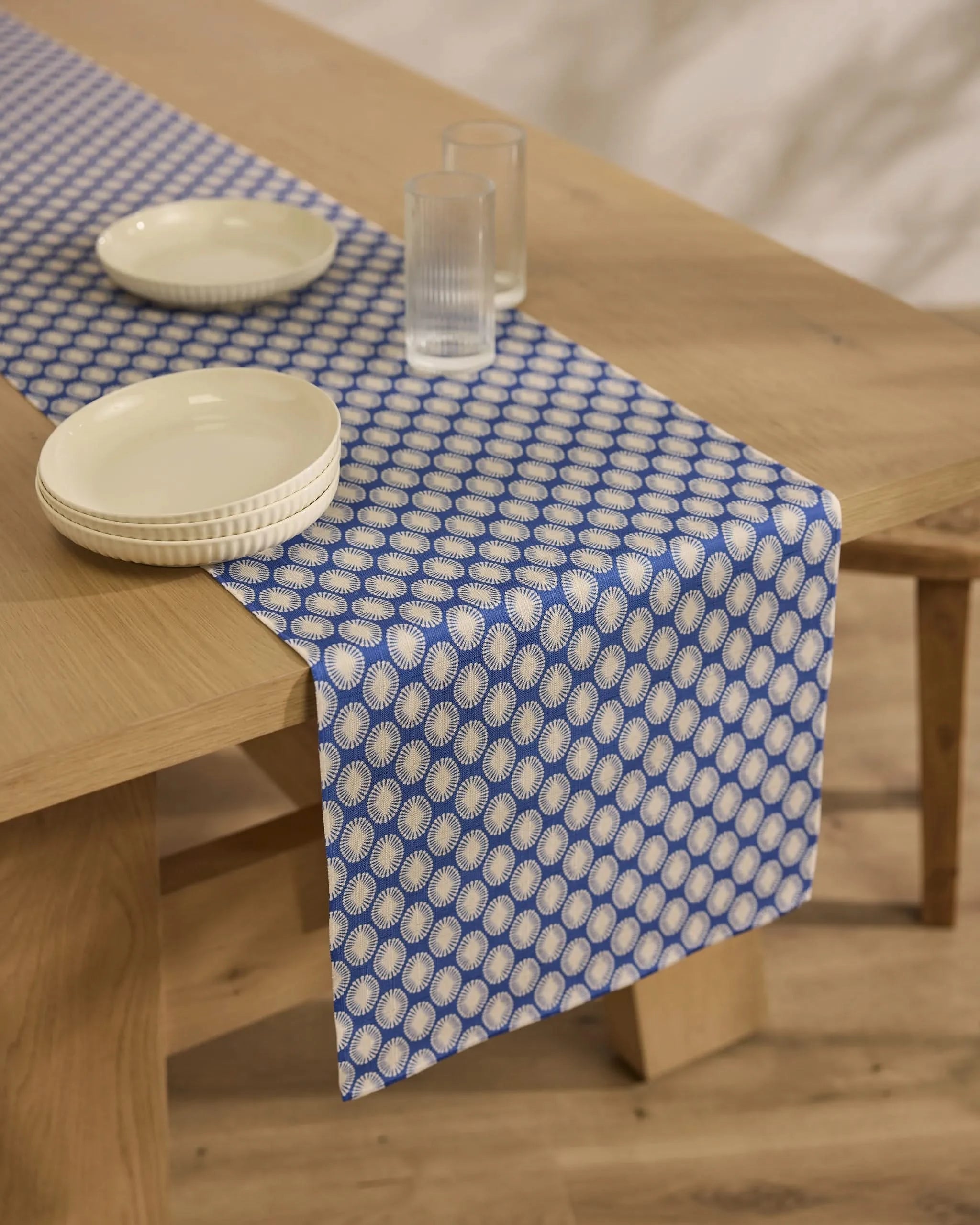 A minimal, modern setting featuring the Lagoon stain-resistant table runner by Geometry in blue and white, styled on a wooden dining table with two white plates and two ribbed drinking glasses.