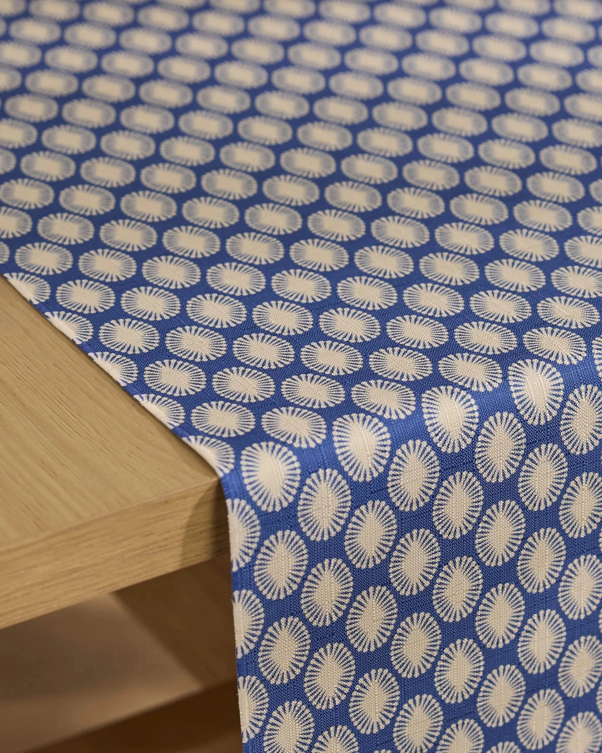 The Geometry Lagoon table runner, featuring a blue stain-resistant fabric with white sunburst patterns, is draped over the edge of a light wooden table.