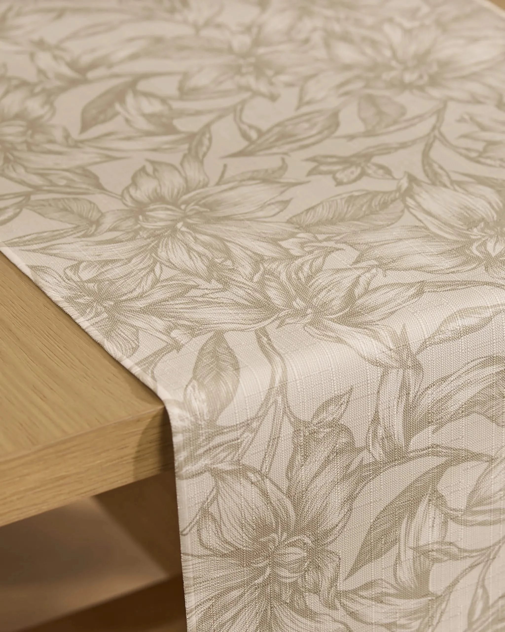 The Alba table runner by Geometry, crafted from recycled materials, features a subtle floral pattern in light beige tones with delicate flower and leaf designs, elegantly displayed over a wooden table.