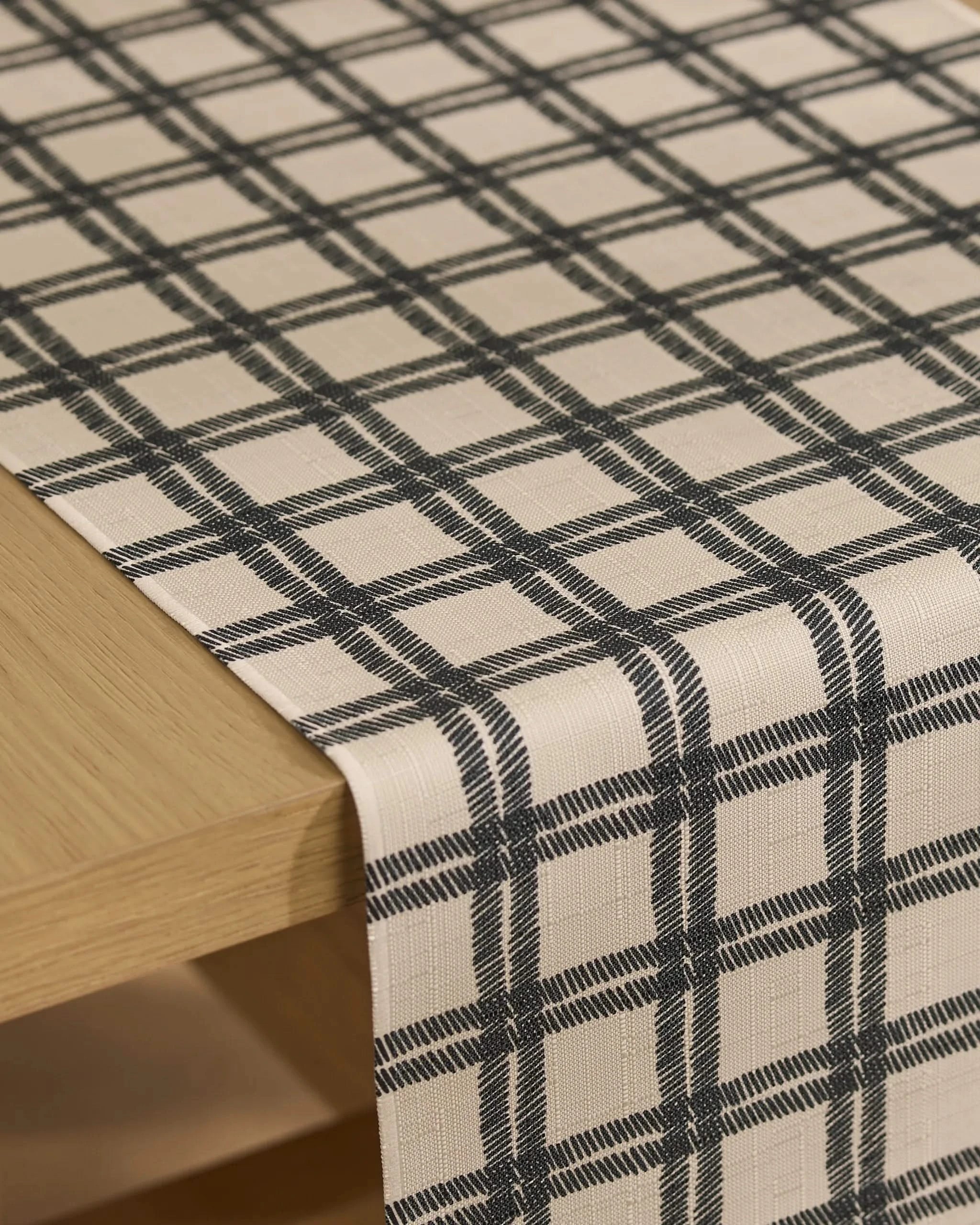 The Geometry Blackstone table runner, made from recycled fabric, features a beige base with a textured finish and a black plaid pattern of evenly spaced horizontal and vertical lines draped over a light wooden table.