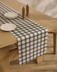 A Blackstone dining table by Geometry features a stain-resistant black and white plaid table runner, three white plates, and two bronze salt and pepper shakers, set on a light wooden floor.