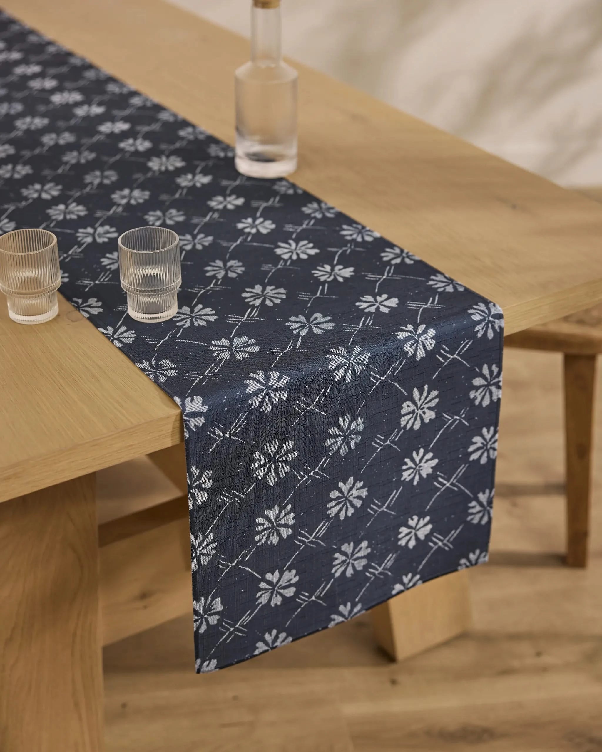 The Geometry Bonnie Indigo table runner, featuring a white floral pattern on dark blue, is stain-resistant and set on a wooden dining table with two clear glasses and a glass bottle. The floor is light wood.