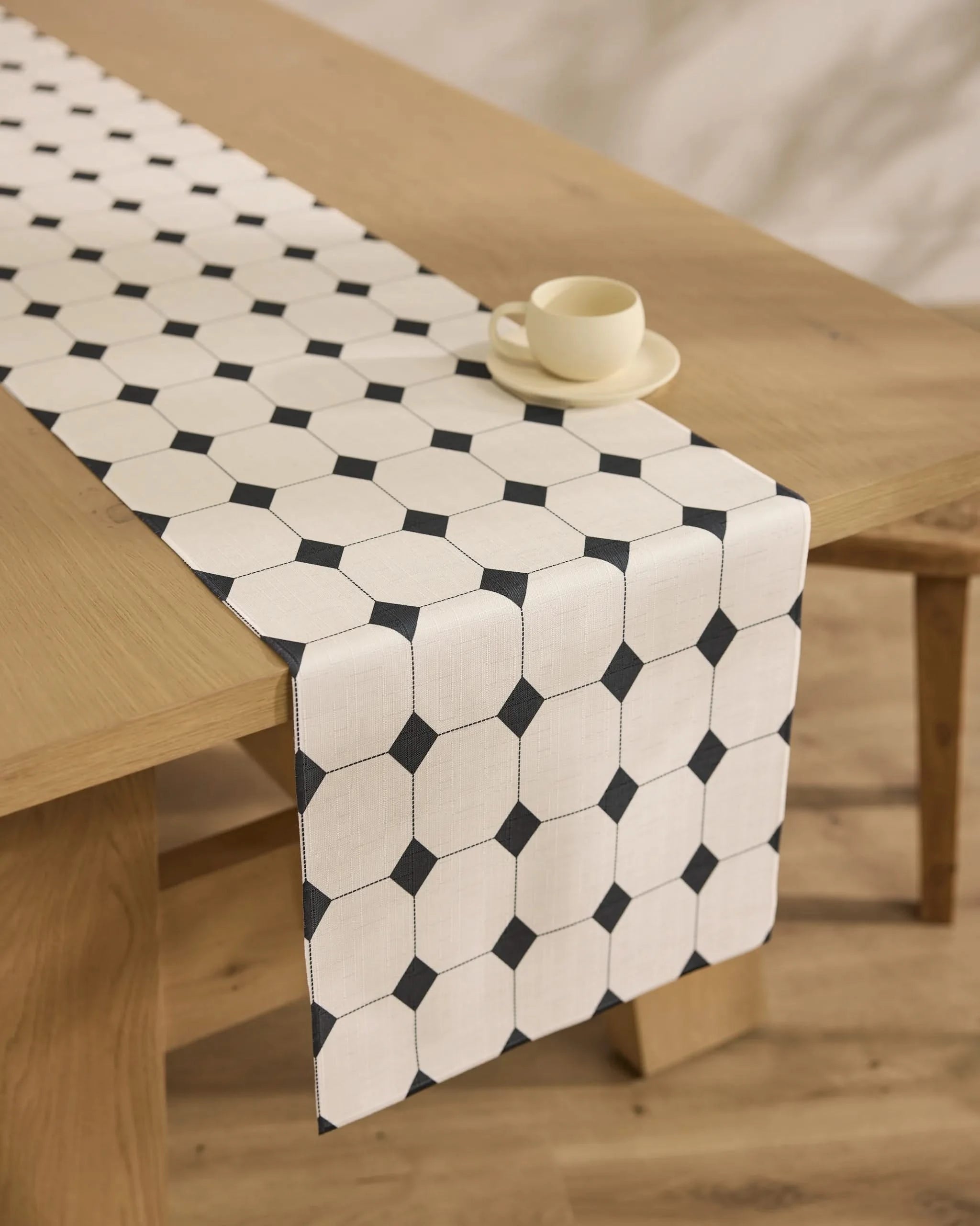 A wooden table with a Geometry Espresso Mosaic table runner in black and white; a cream-colored cup and saucer rest on top.