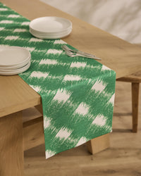 A light wood dining table features Geometrys Hairy Check Green stain-resistant runner, four stacked white plates, and two forks on top, set on a wooden floor.