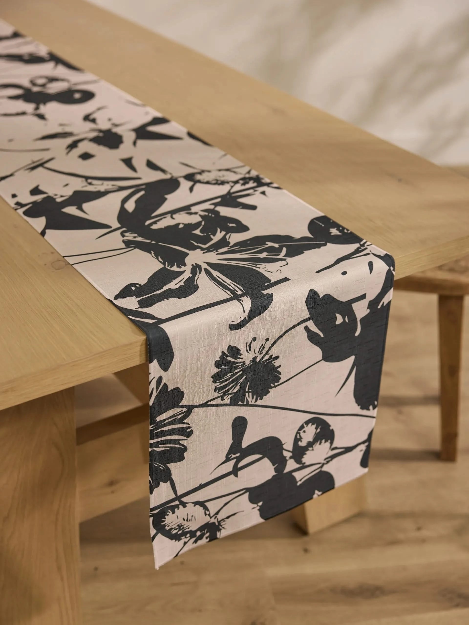 The Inverted Floral table runner by Geometry, featuring a bold black floral print on beige and stain-resistant fabric, is elegantly displayed on a light wood table in a minimalist room with wooden floors and neutral walls.