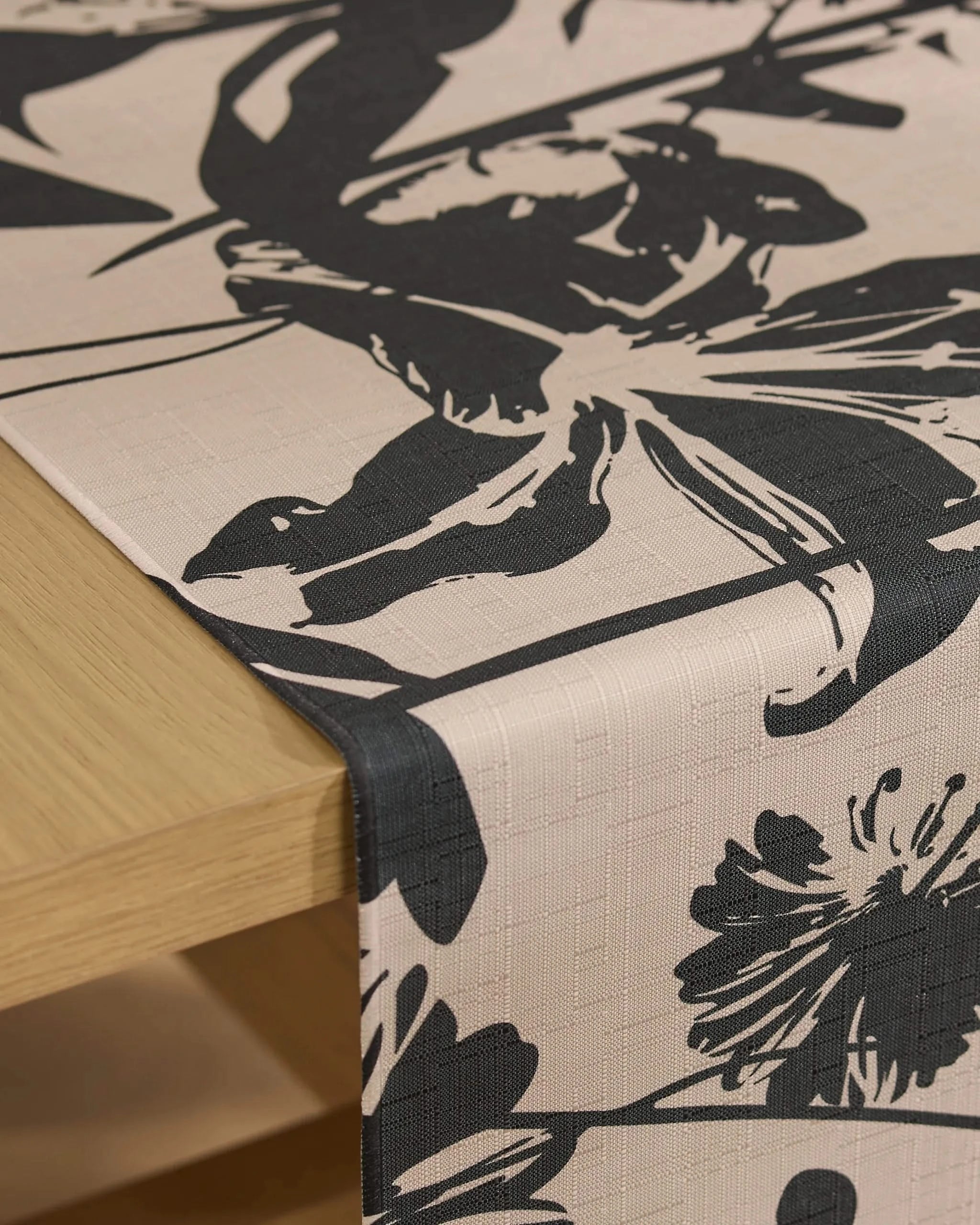 The Geometry Inverted Floral table runner, in beige with a bold black floral pattern and stain-resistant fabric, is draped over the edge of a light wooden table.
