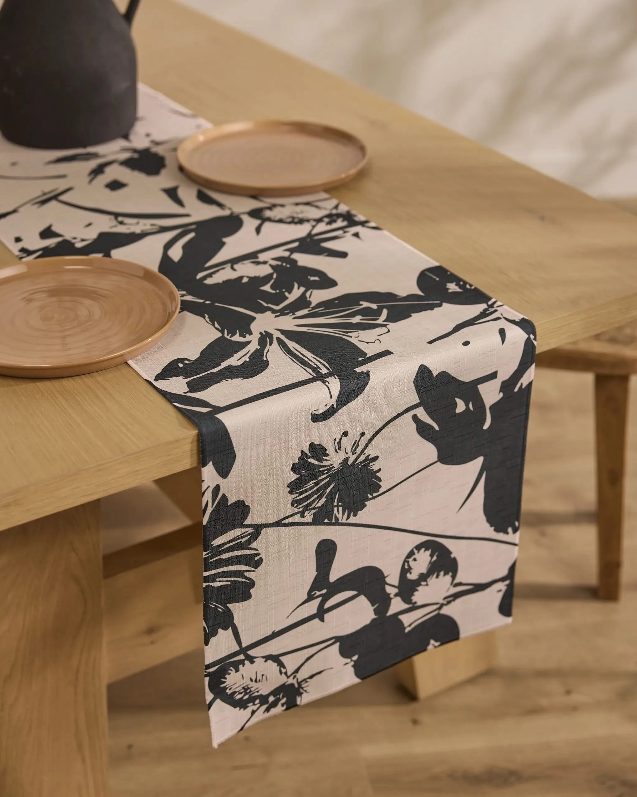 A wooden dining table with a stain-resistant Geometry Inverted Floral table runner, set with simple round wooden plates and a dark pitcher.