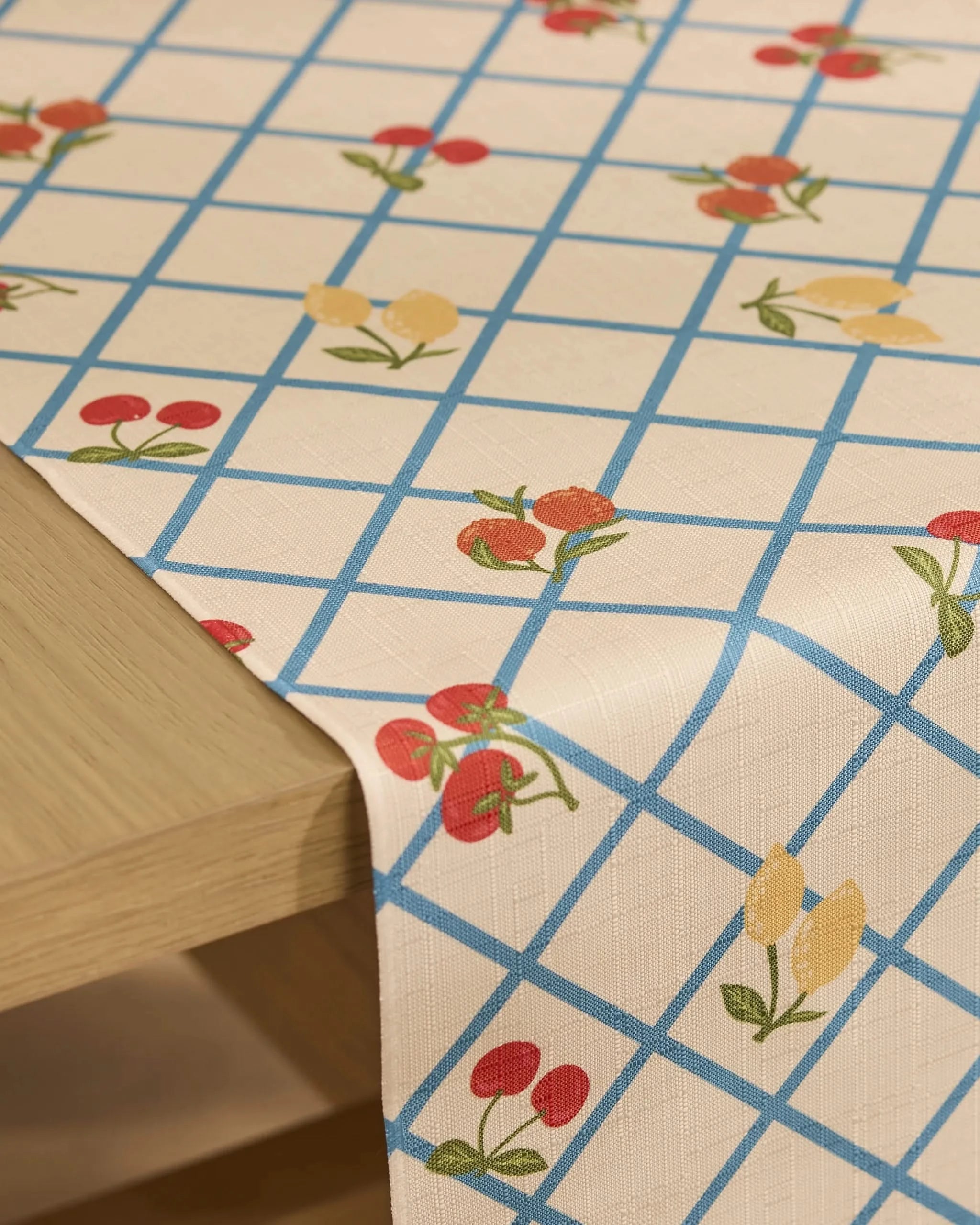 A close-up of a wooden table with Geometrys Italian Summer Grid table runner, featuring a blue grid and small red and yellow fruit with green leaves. Made from recycled materials, the runner drapes elegantly over the edge.