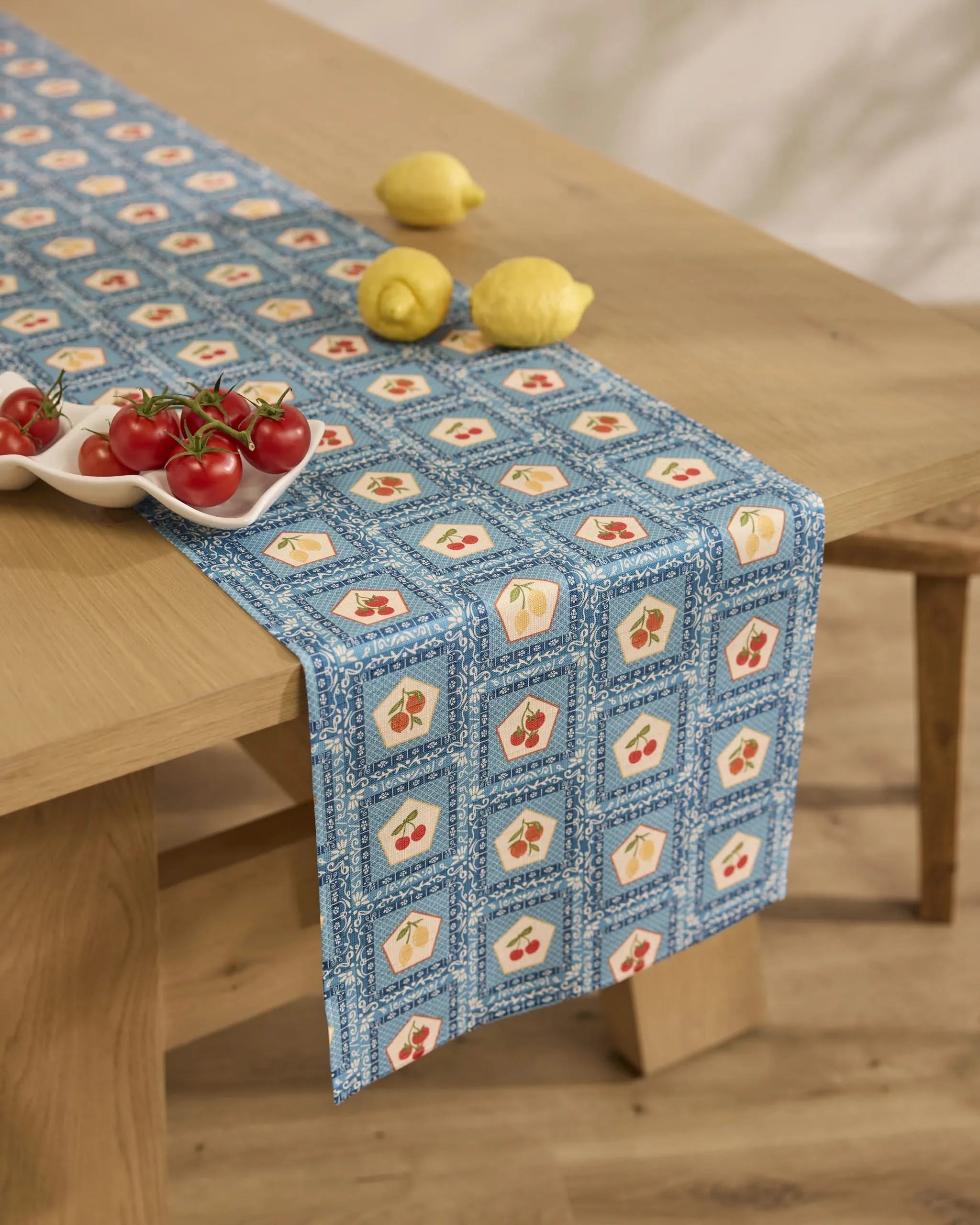 A wooden table with a Geometry Italian Tiles table runner, featuring blue tile designs, holds a ceramic dish of tomatoes and two lemons. The scene is set in a bright room with a neutral background.