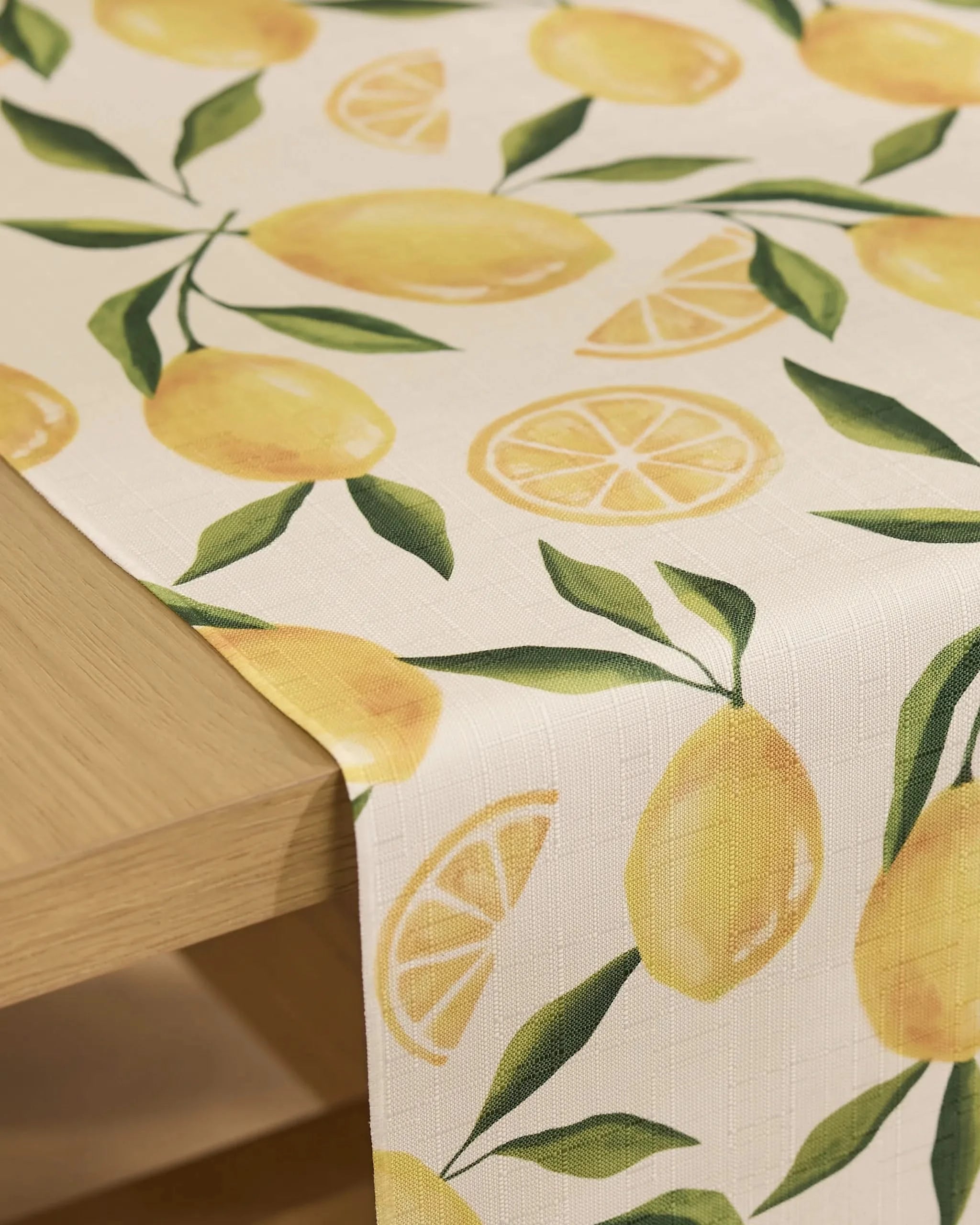 The Geometry Limoncello table runner features a cheerful, stain-resistant design with whole lemons, lemon slices, and green leaves, draping stylishly over a light wooden table—perfect for practical, everyday use.