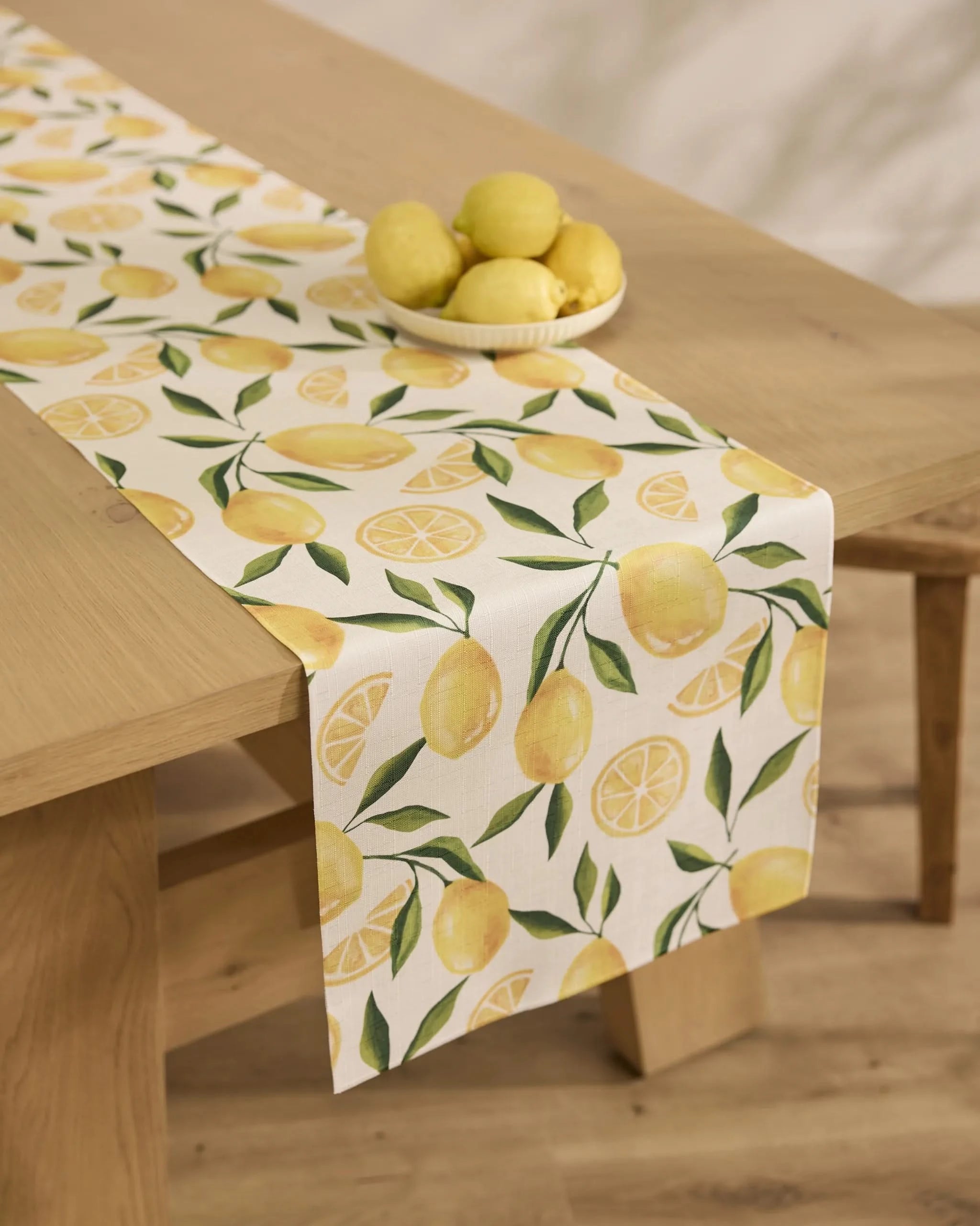 A wooden table with a Geometry Limoncello stain-resistant runner, featuring yellow lemons and green leaves on white, topped with a bowl of whole lemons.