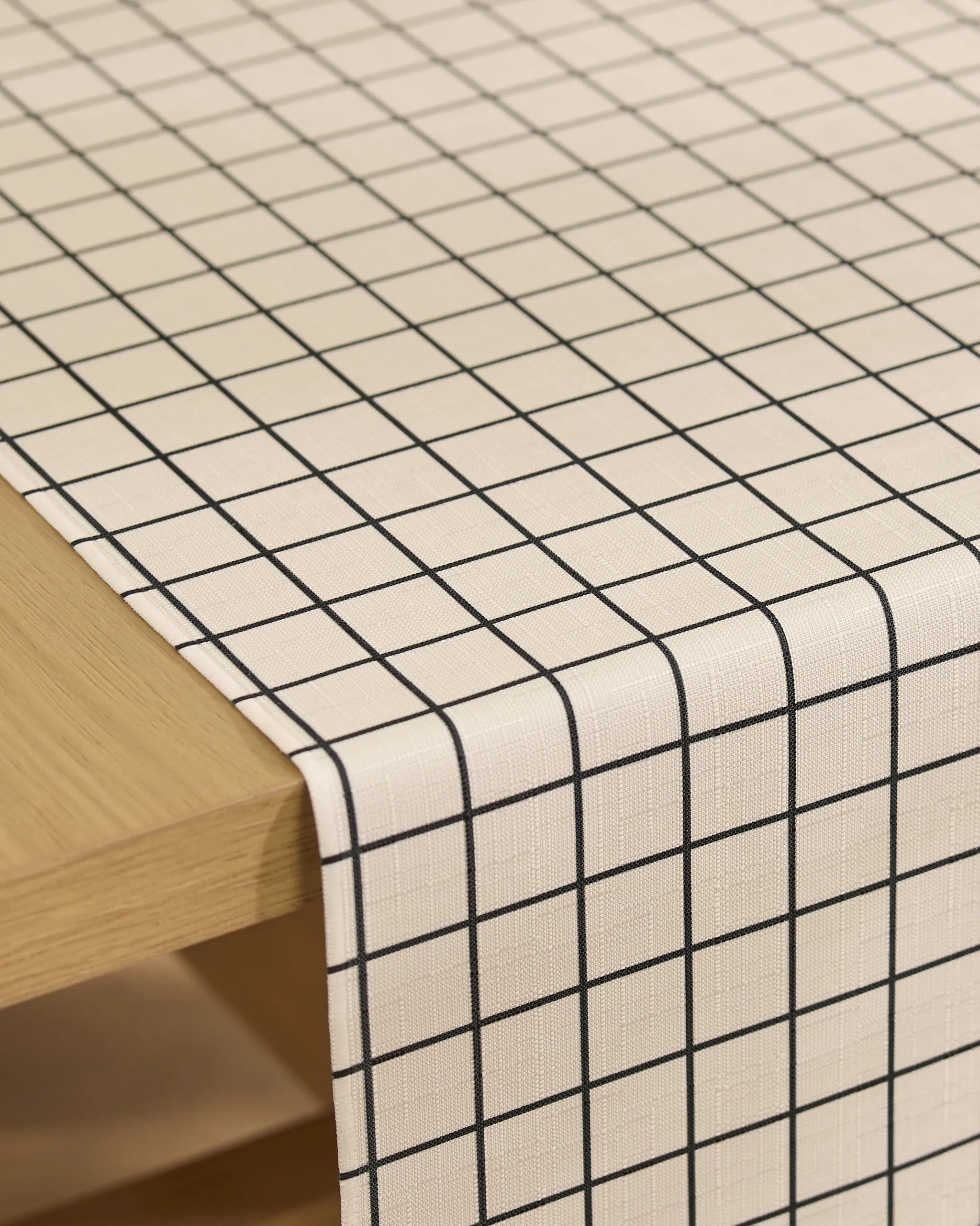 The Geometry Metro Grid table runner, featuring a stain-resistant black grid pattern on a white background, is neatly draped over the edge of a light wooden table.