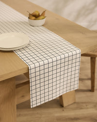 A light wooden table is topped with Geometry’s Metro Grid stain-resistant runner, showcasing its black grid design. Three white plates are stacked next to a small bowl of pears.