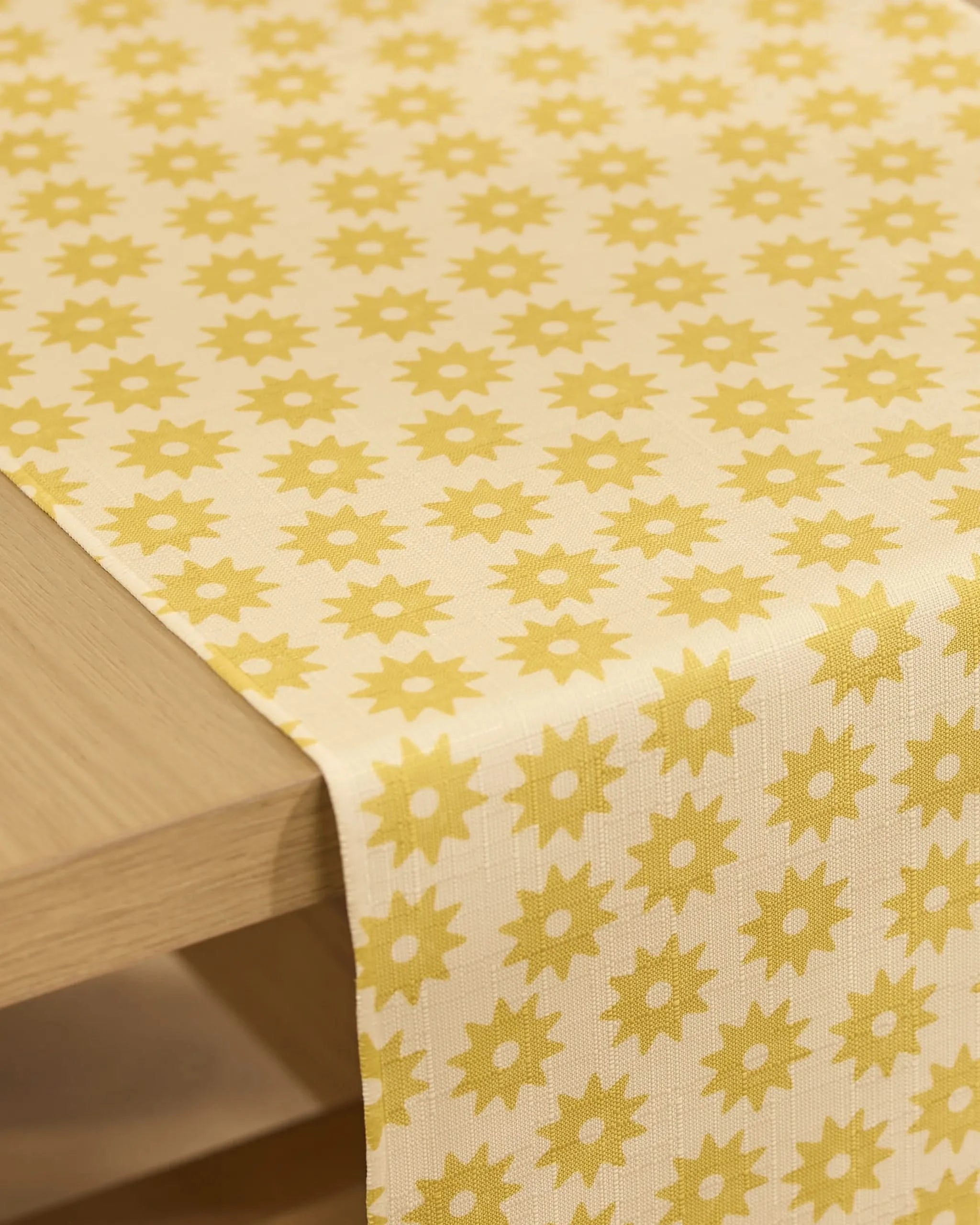 The Geometry Sunny Sun stain-resistant table runner, featuring a repeating yellow floral pattern on a cream background, drapes over the edge of a light wooden table.