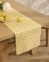 A light wood table features Geometry’s Sunny Sun yellow and white floral-patterned, stain-resistant table runner, topped with a bowl of lemons, a single lemon, and a white flower for a fresh, summery vibe.