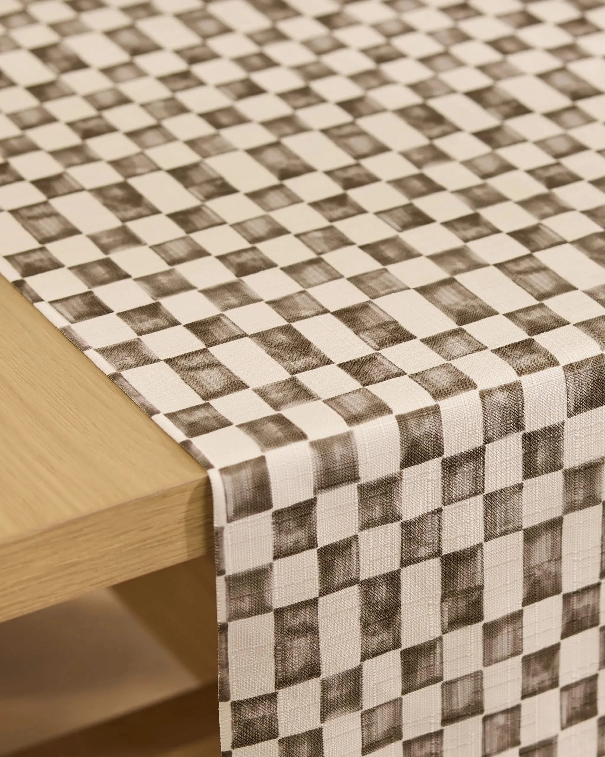 A close-up of a light wooden table with the Tavern table runner by Geometry in white and gray checks, featuring a textured, brushed pattern. The versatile, stain-resistant runner drapes neatly over the edge.