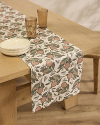 The Geometry Valencia stain-resistant table runner features an orange fruit and green leaf pattern. Two stacked white bowls and two brown glasses sit on it atop a wooden table with a light-colored wooden floor.