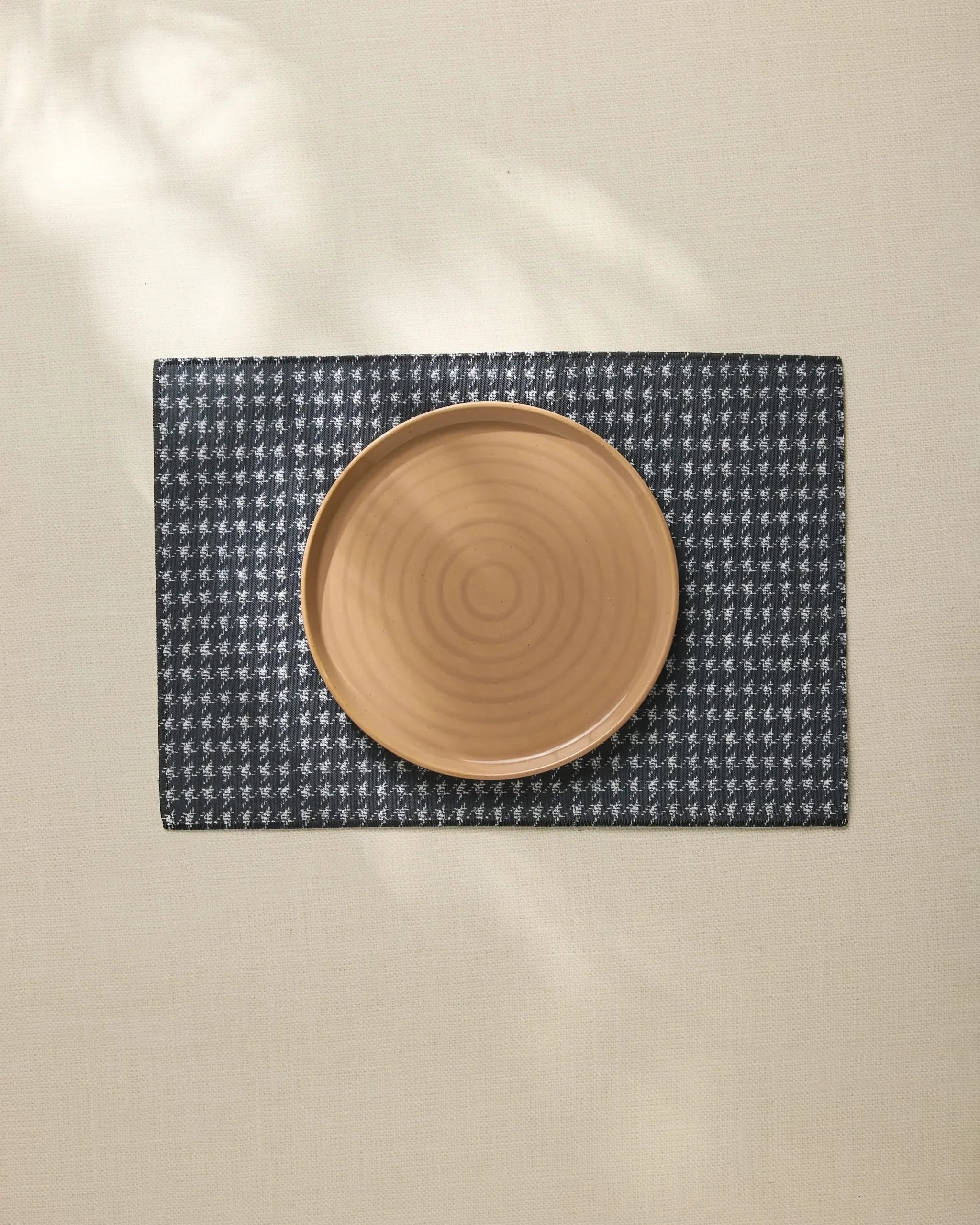 A round, light brown ceramic plate is centered on the Geometry Houndstooth Black placemat, made from recycled materials and set atop a beige tablecloth. Photo is taken from above in soft natural light.