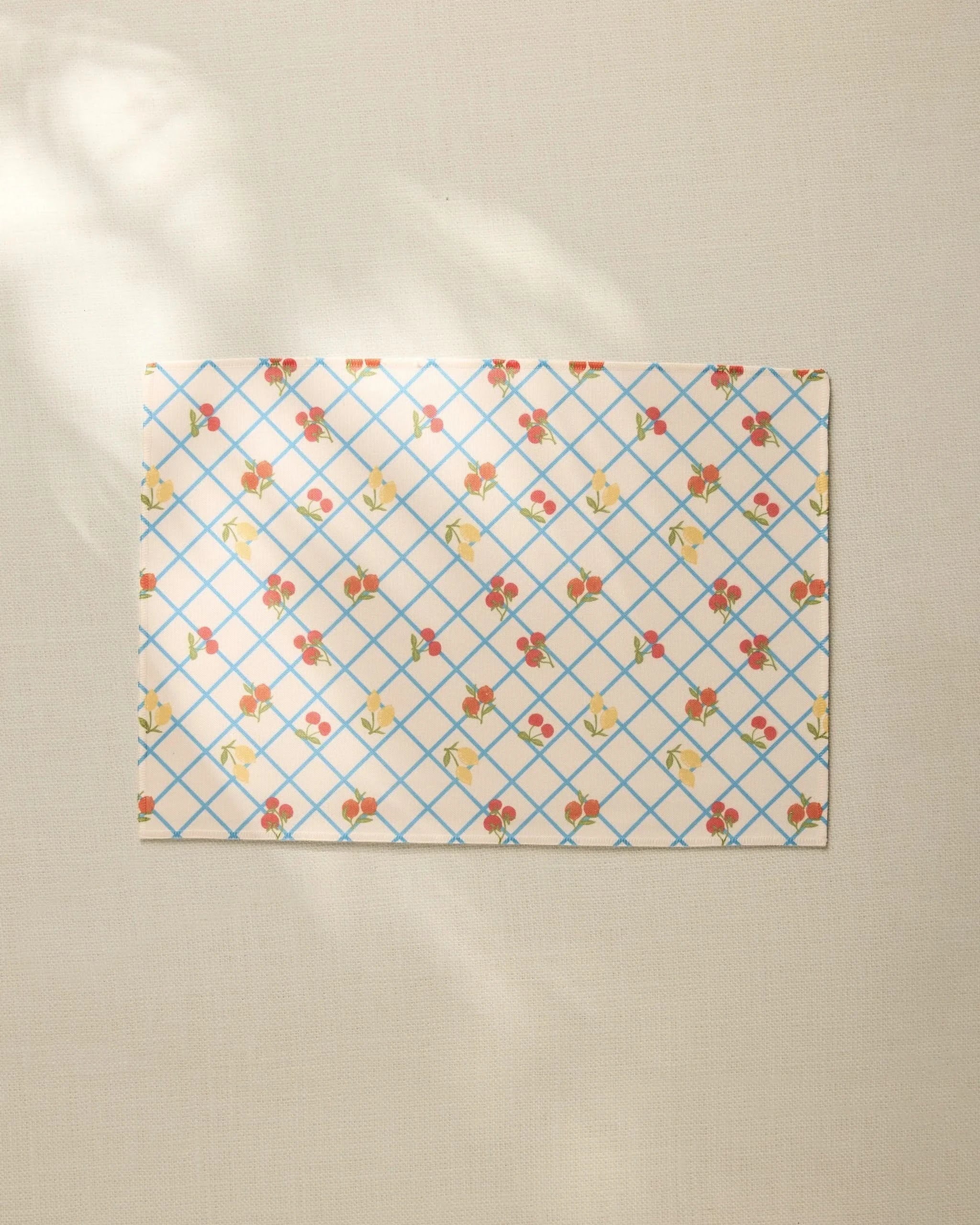 The Italian Summer Grid by Geometry, a rectangular sheet with a blue grid and small floral pattern, rests on a beige textured surface, its recycled placemat style highlighted by soft sunlight and gentle shadows.