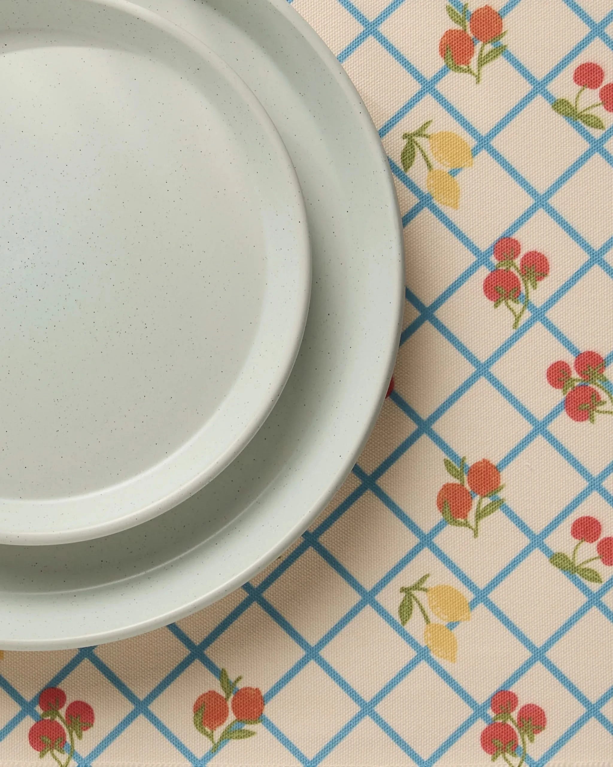 Two white ceramic plates stacked on a table with Geometry’s Italian Summer Grid blue-grid tablecloth adorned with red and yellow fruit patterns, complemented by machine washable placemats for easy cleanup.