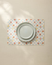A plain light gray plate sits on the Geometry Italian Summer Grid Placemat, featuring a blue grid and small red and yellow floral pattern, atop a neutral tablecloth.