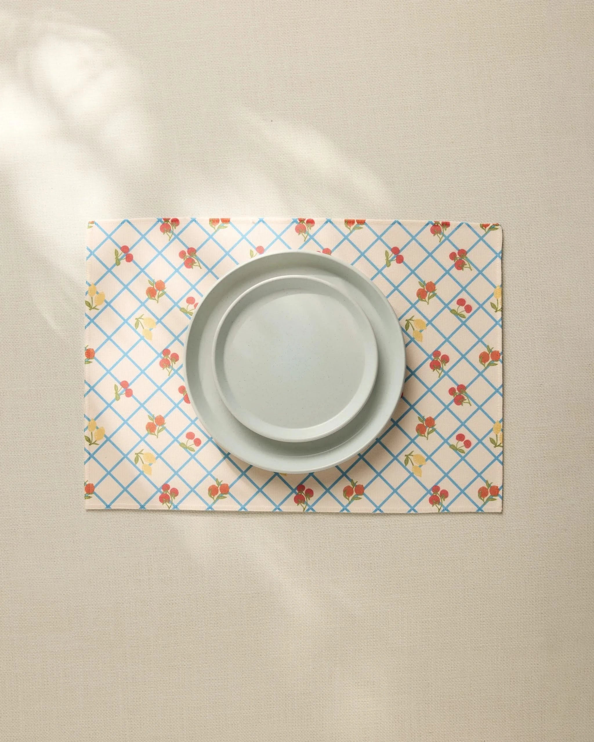 A plain light gray plate sits on the Geometry Italian Summer Grid Placemat, featuring a blue grid and small red and yellow floral pattern, atop a neutral tablecloth.