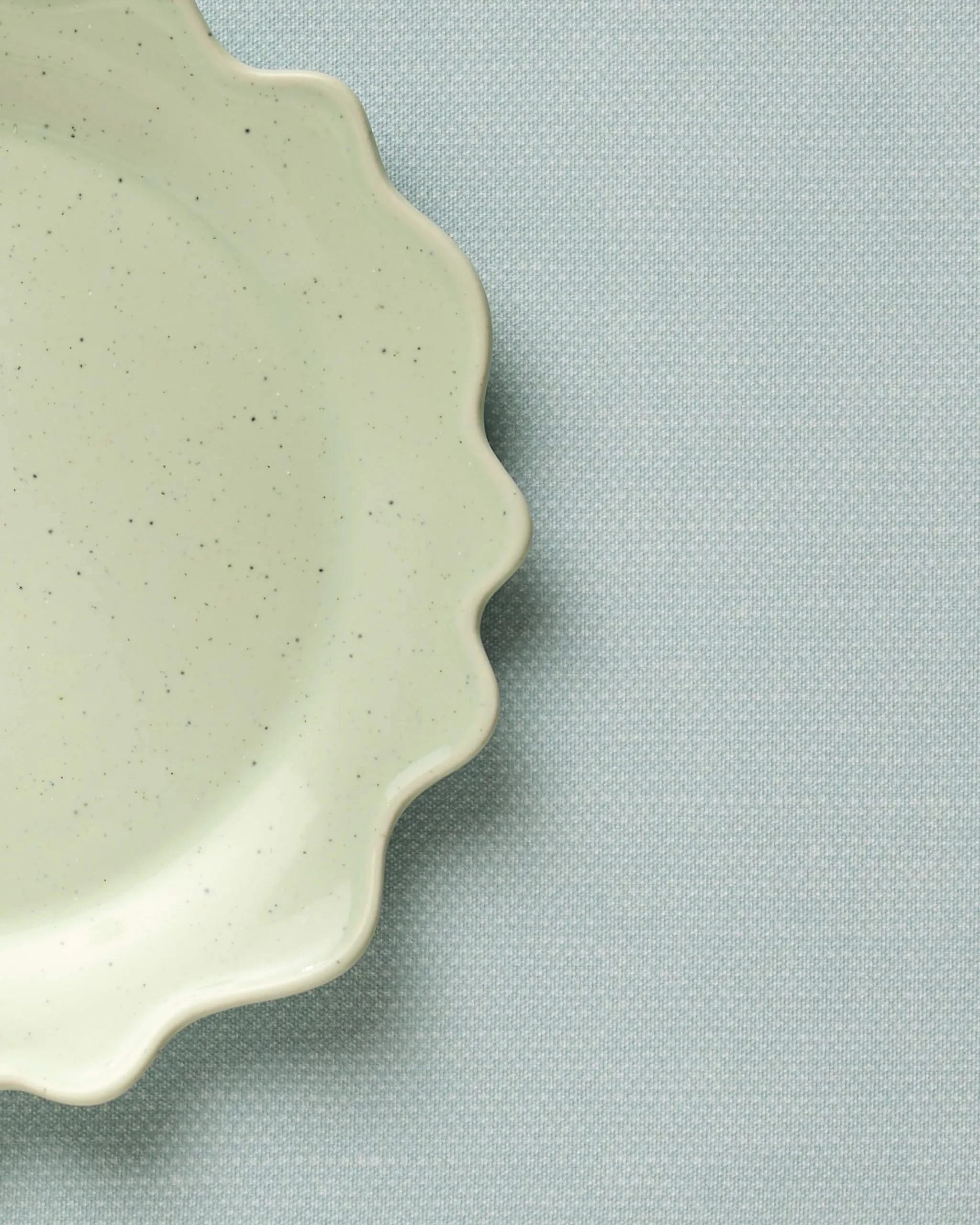 A close-up shows a pale green, speckled ceramic plate with a wavy edge resting on a textured Geometry Chambray Light Blue placemat. Only part of the plate appears in the upper left corner.