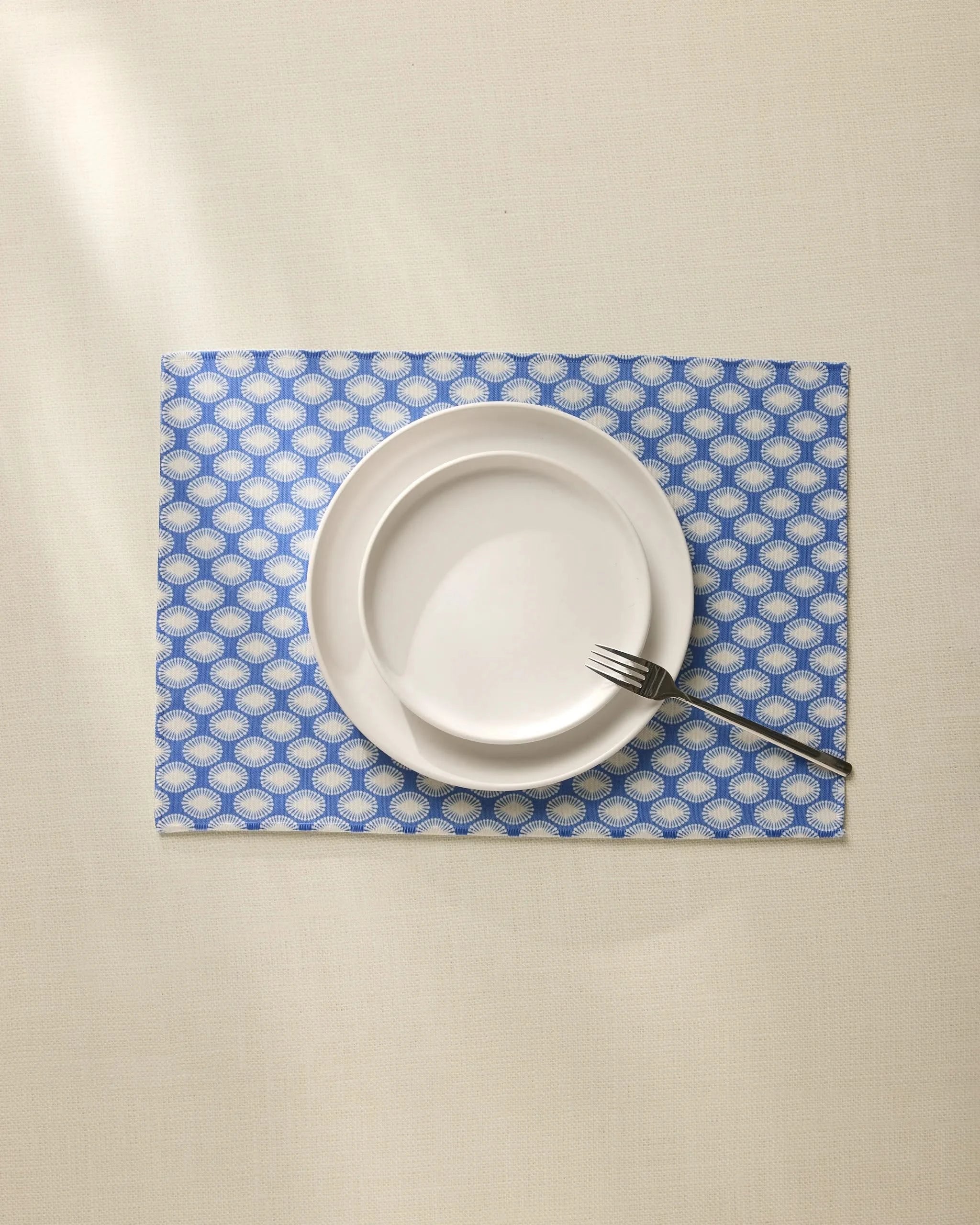 A white plate and small bowl stacked with a fork on the edge sit on a cream tablecloth over stylish Geometry Lagoon Placemats in blue and white patterns.
