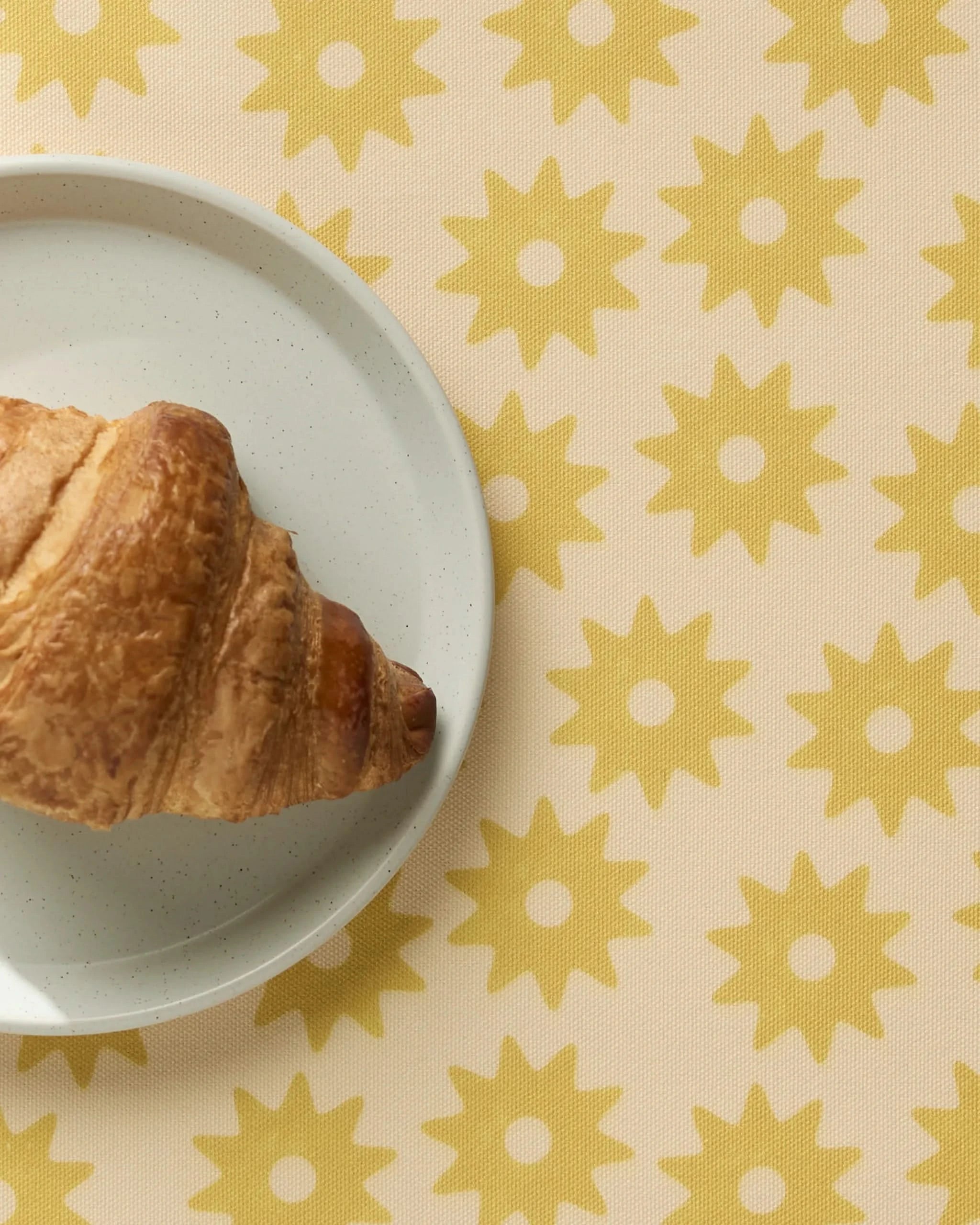 A croissant on a white plate rests on machine washable Geometry Sunny Sun placemats atop a beige tablecloth decorated with yellow sun-like patterns.