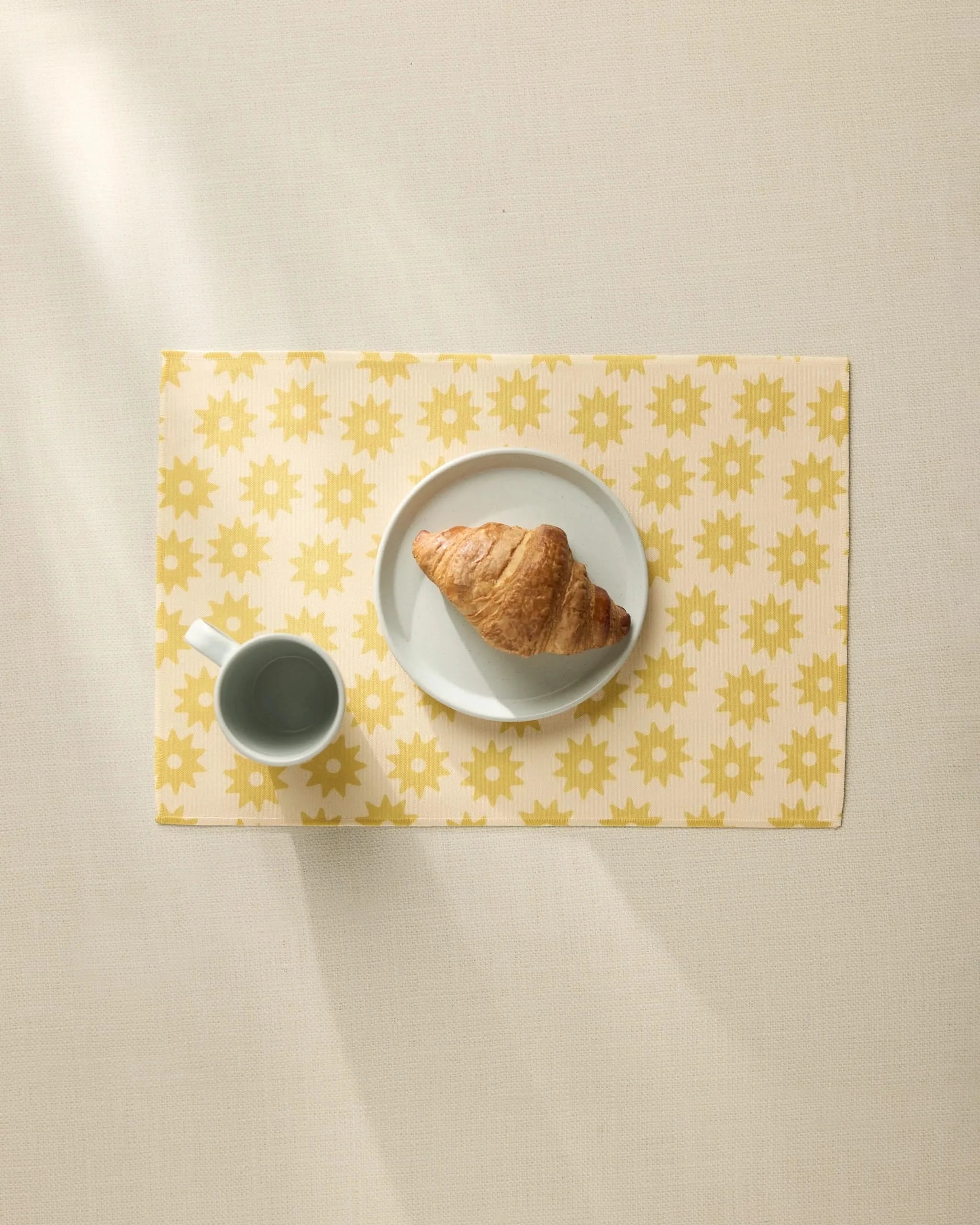 A croissant on a white plate and a cup of coffee rest on the Geometry Sunny Sun placemat, featuring a yellow sun pattern, viewed from above on a light beige table.