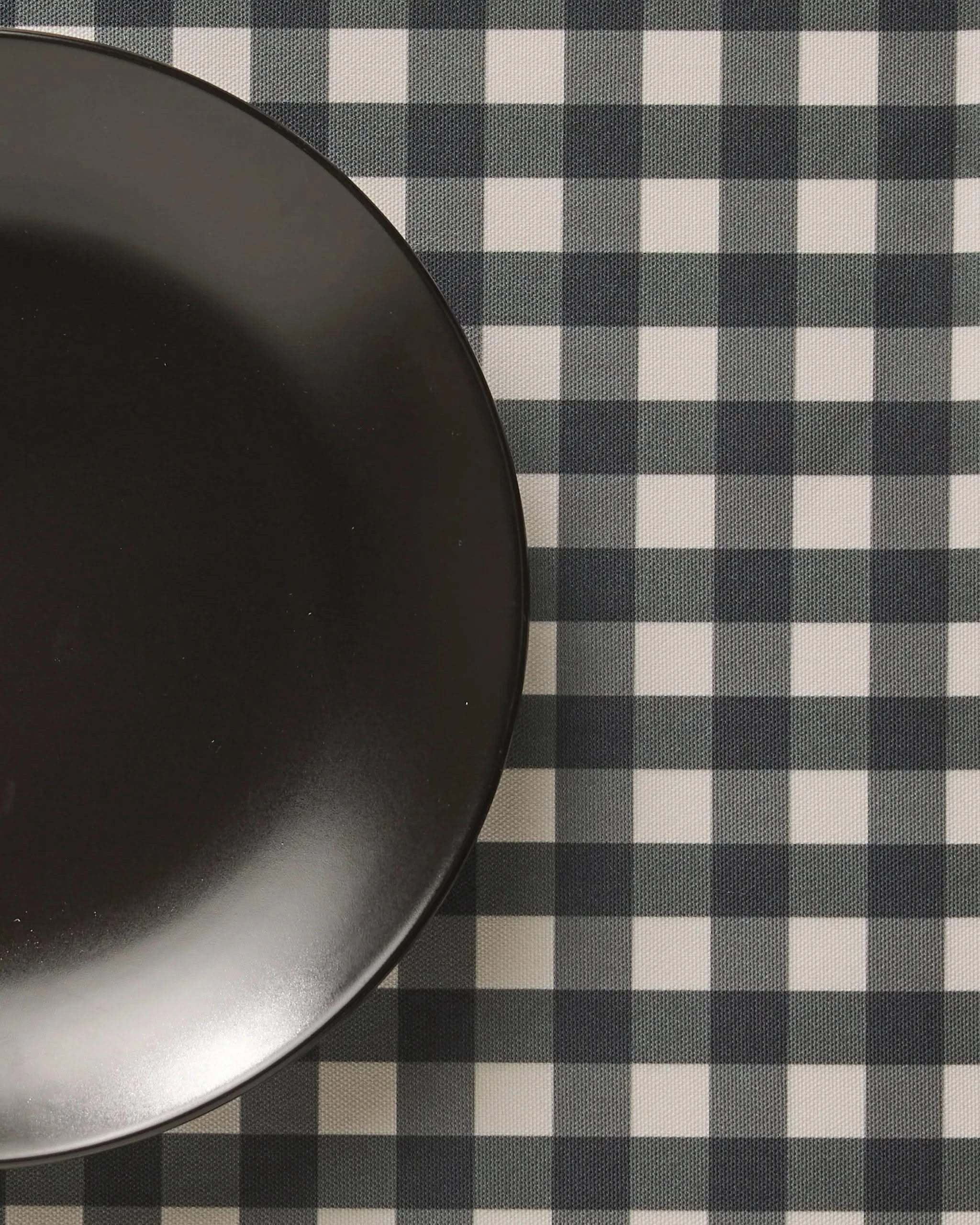 A black plate sits on the left side of a checkered tablecloth with a black and white Charcoal Gingham pattern from Geometry, featuring stylish, machine washable placemats for easy care.