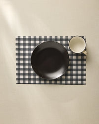 A black bowl and white mug rest on the Charcoal Gingham placemat by Geometry, neatly arranged atop a light-colored tablecloth.