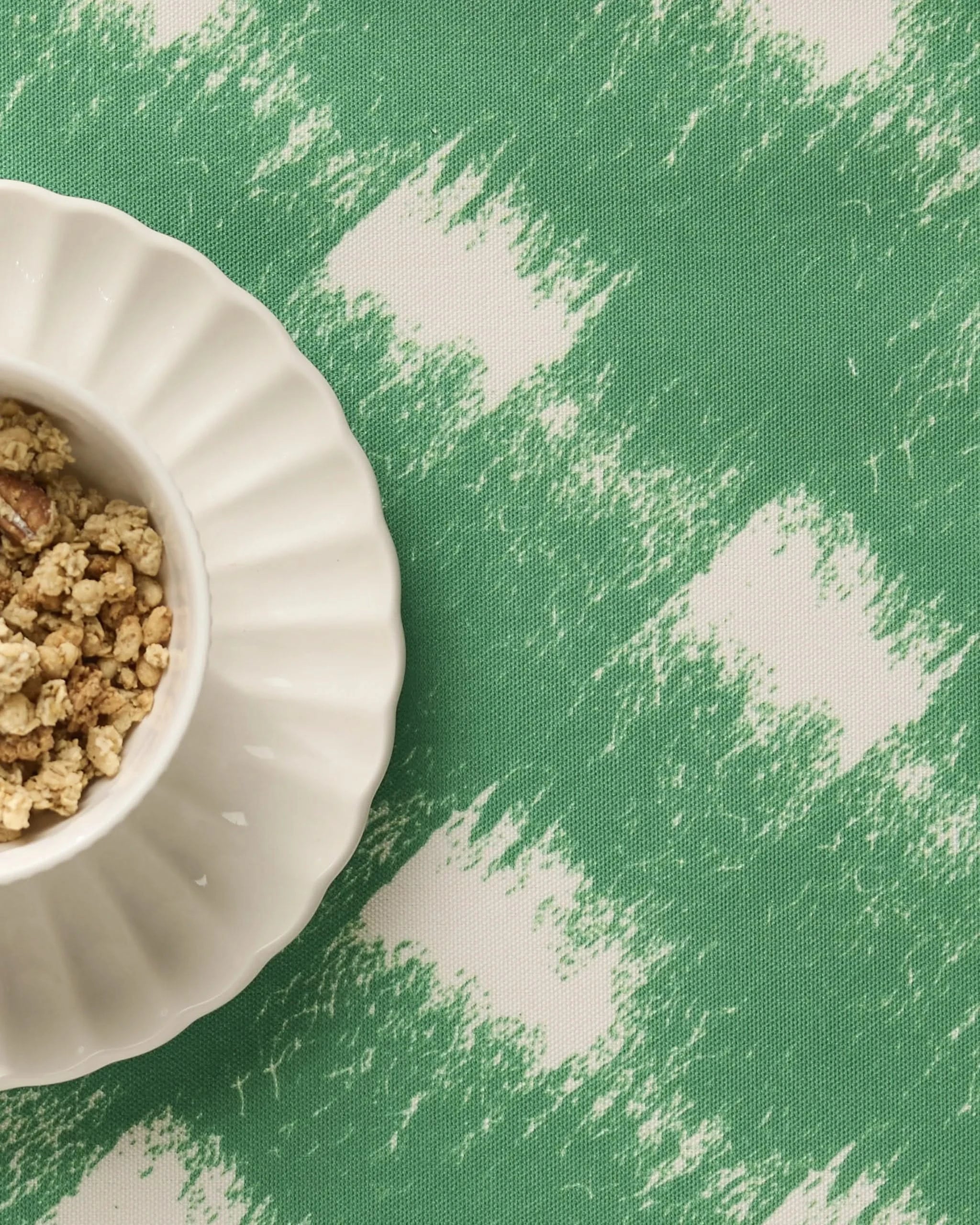 A white scalloped bowl of granola sits on Geometrys Hairy Check Green placemat, featuring a green and white brushstroke pattern and a machine-washable surface.