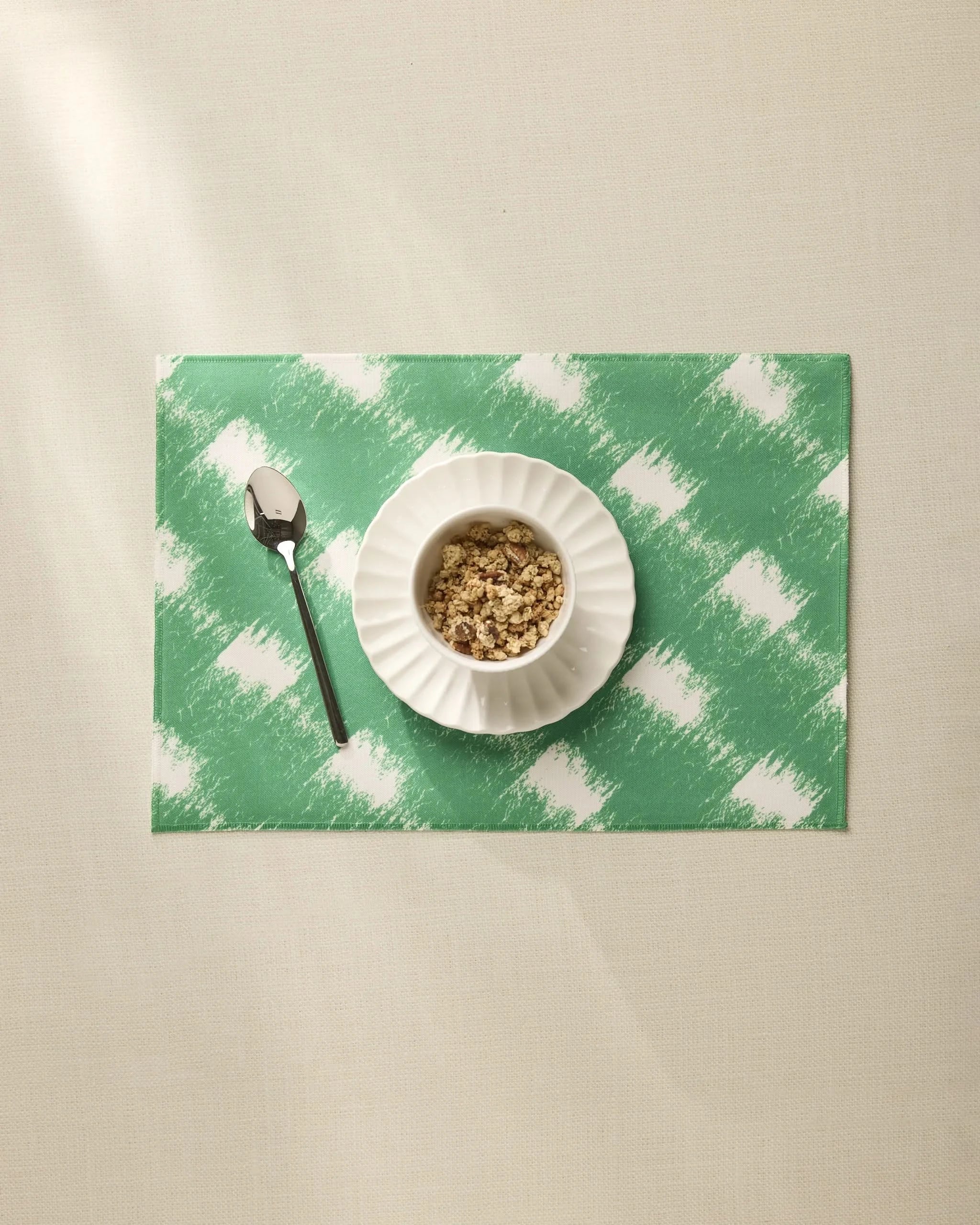 A white bowl of granola rests on the Hairy Check Green placemat by Geometry, with a spoon to the left, all set on a beige tablecloth.