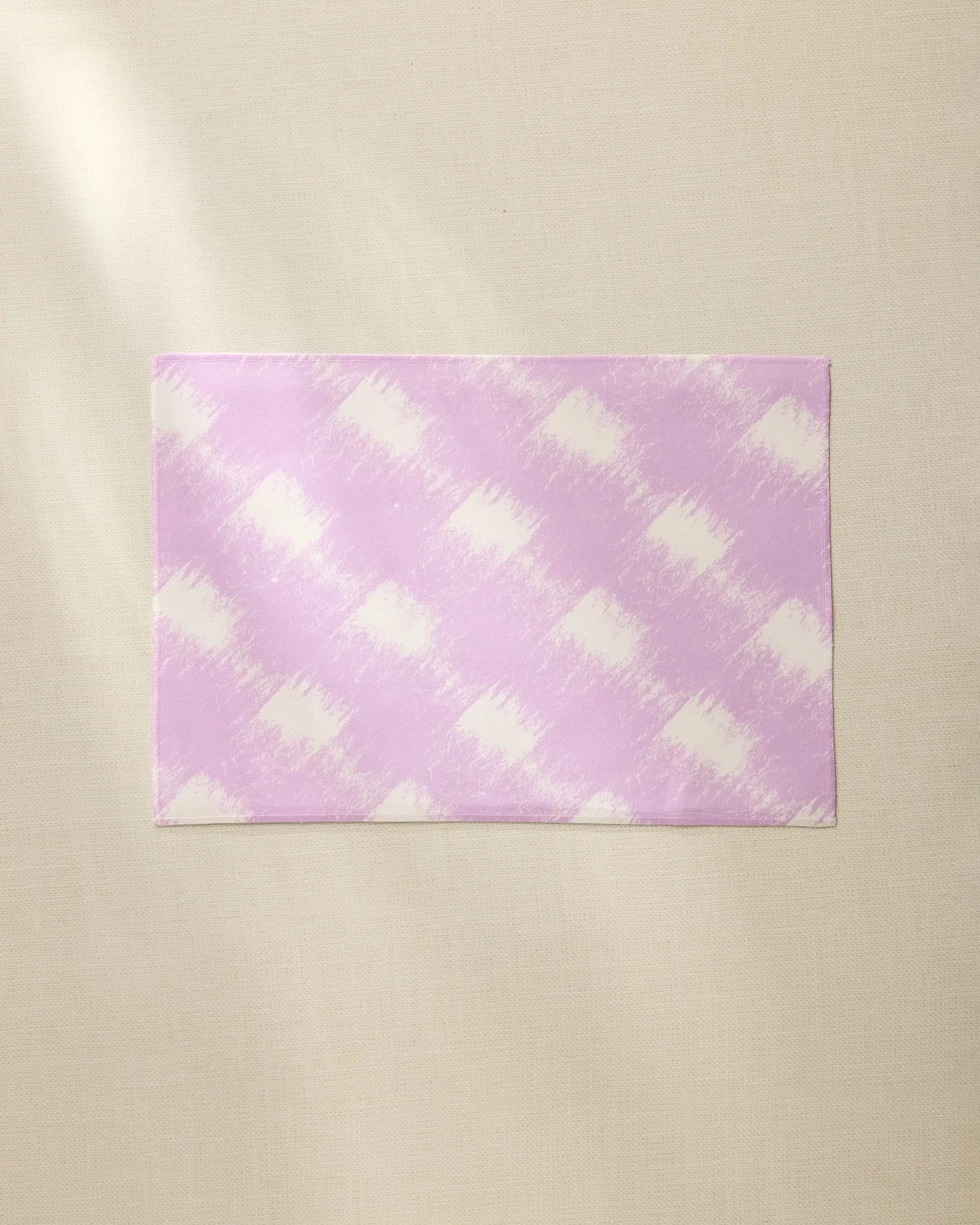 The Geometry Hairy Check Pink placemat features a light purple and white diagonal checkered pattern. Durable and machine washable, its a stylish, practical choice for everyday dining. Shown laid flat on a beige textured surface.