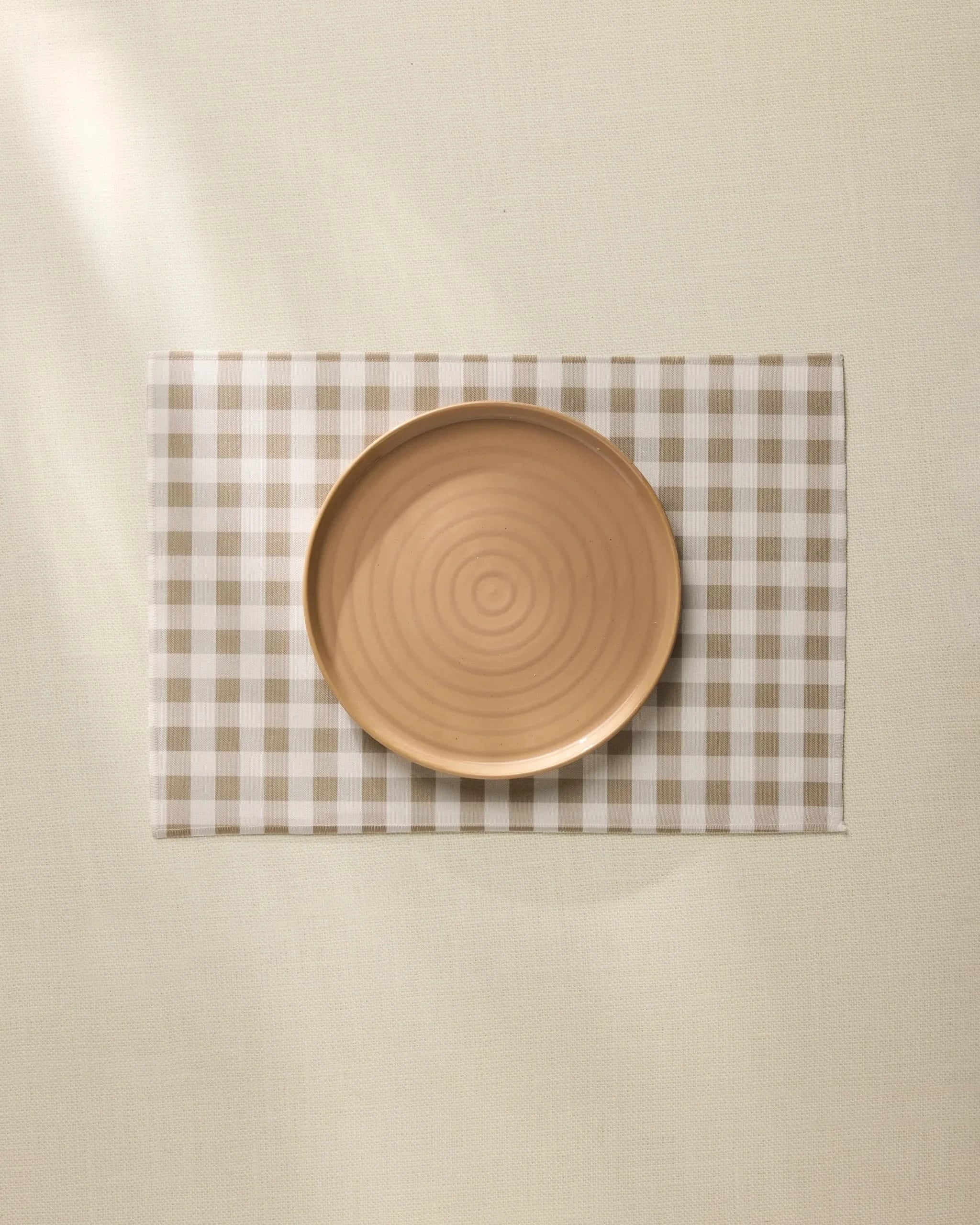 A round, light brown ceramic plate sits centered on the Geometry Hazel Gingham Placemat atop a cream-colored tablecloth.