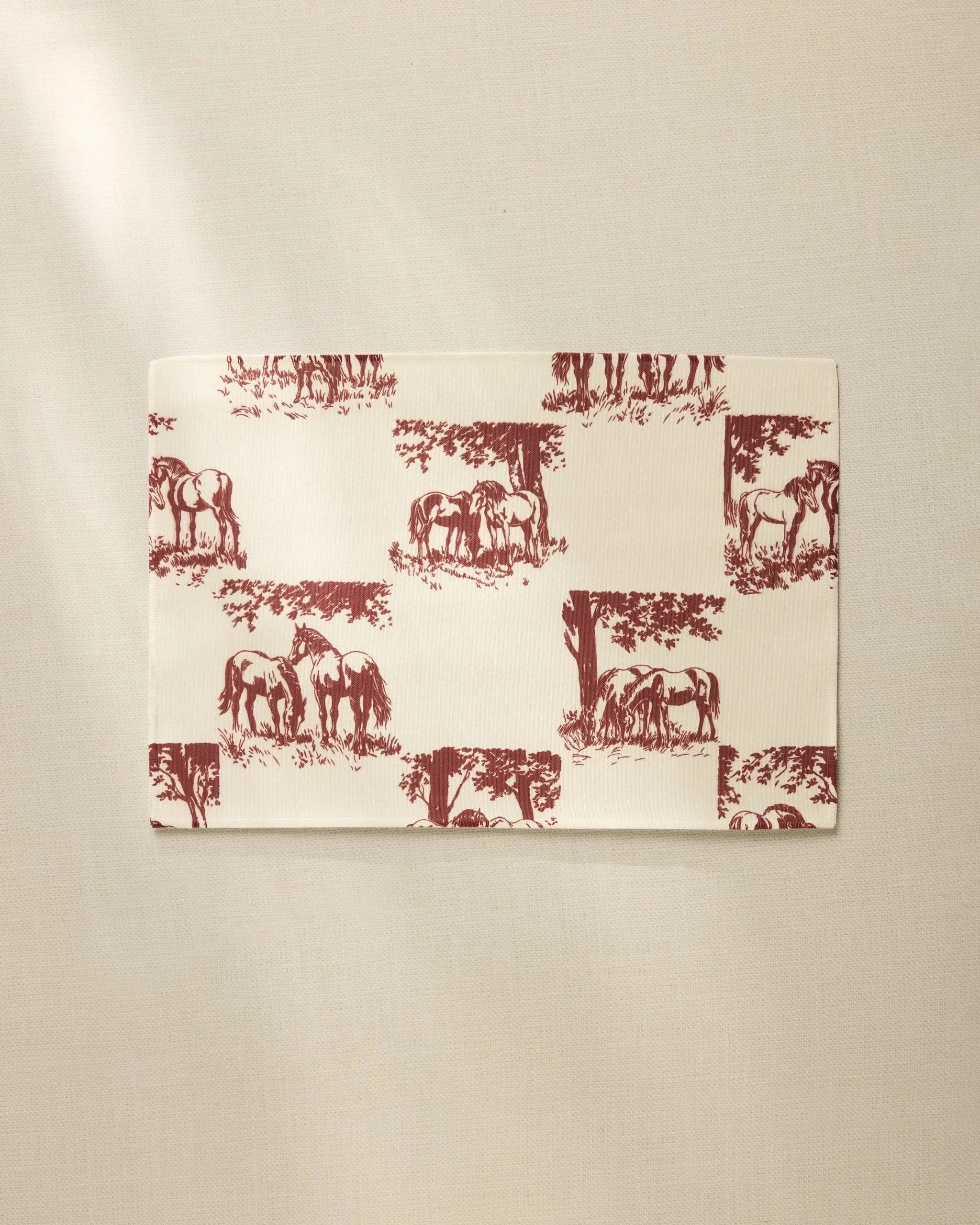The Geometry Horse Check Cream is a rectangular, cream-colored, machine washable placemat featuring maroon horses and trees in a repeating pattern, shown on a neutral, lightly textured surface.