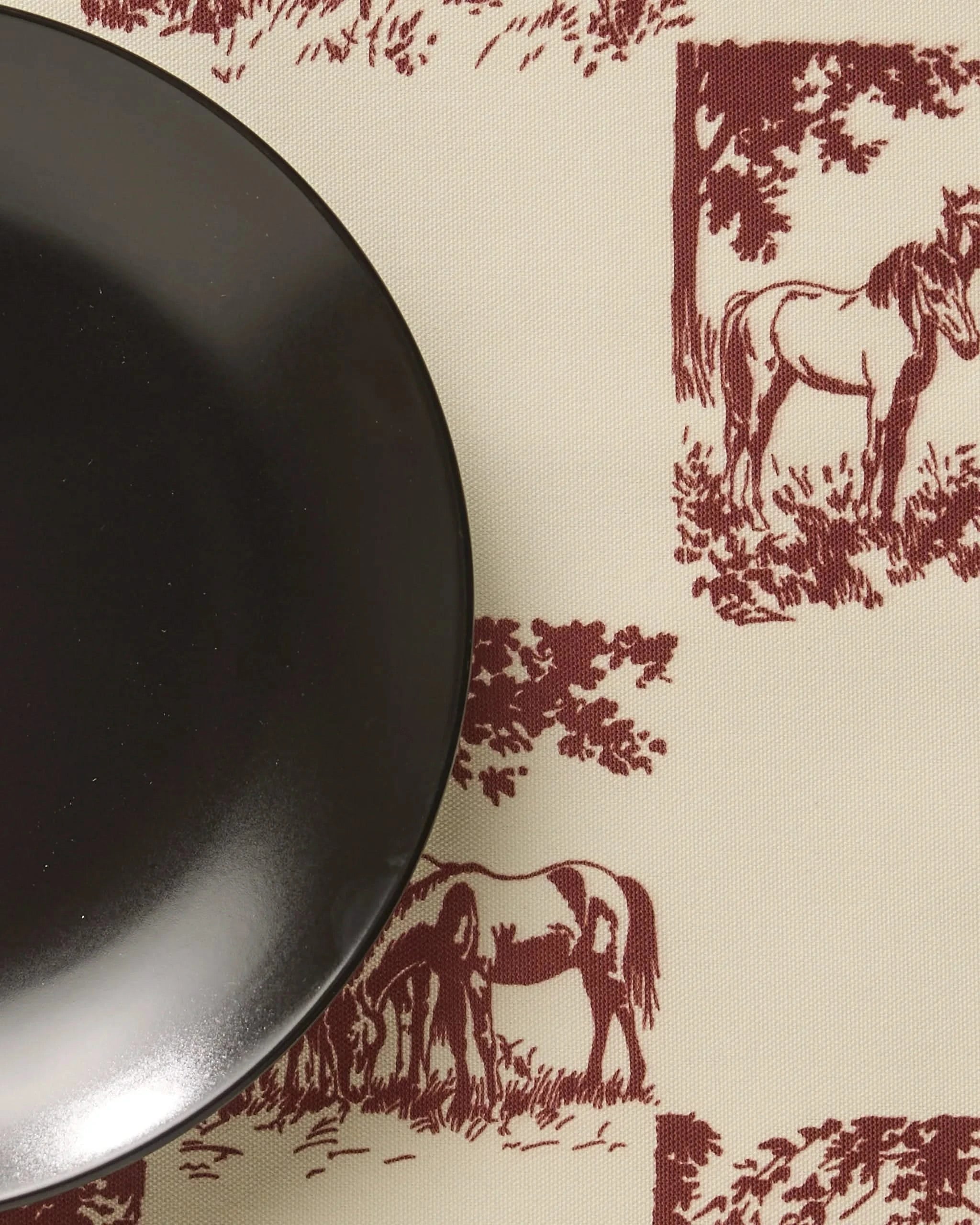 A black plate sits on Geometry’s Horse Check Cream placemat, featuring a cream background with red horse and tree illustrations in a square pattern.