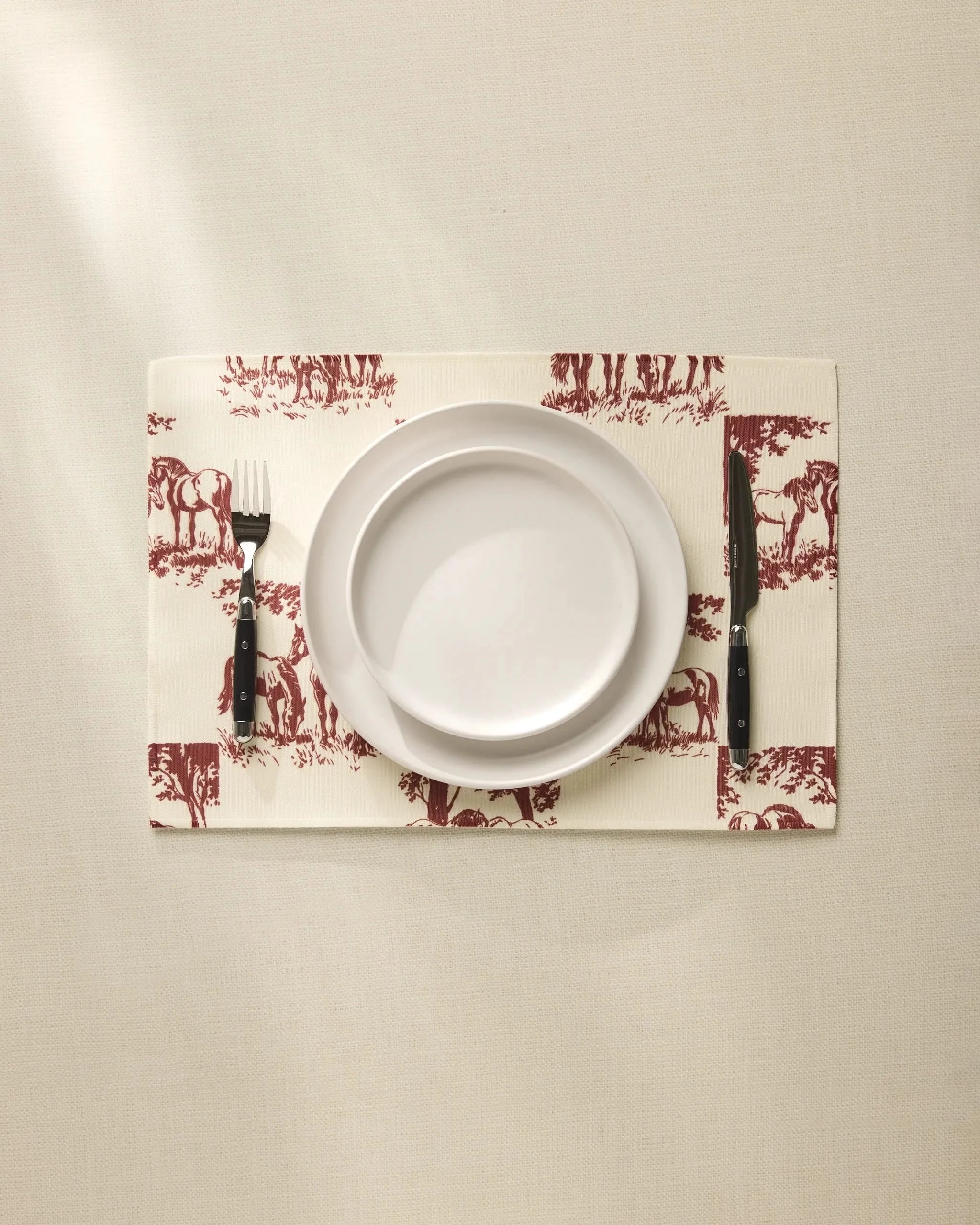 A white plate sits on the Geometry Horse Check Cream placemat with maroon horse designs, flanked by a fork and knife on a beige tablecloth. These machine-washable placemats blend style and easy care for any setting.